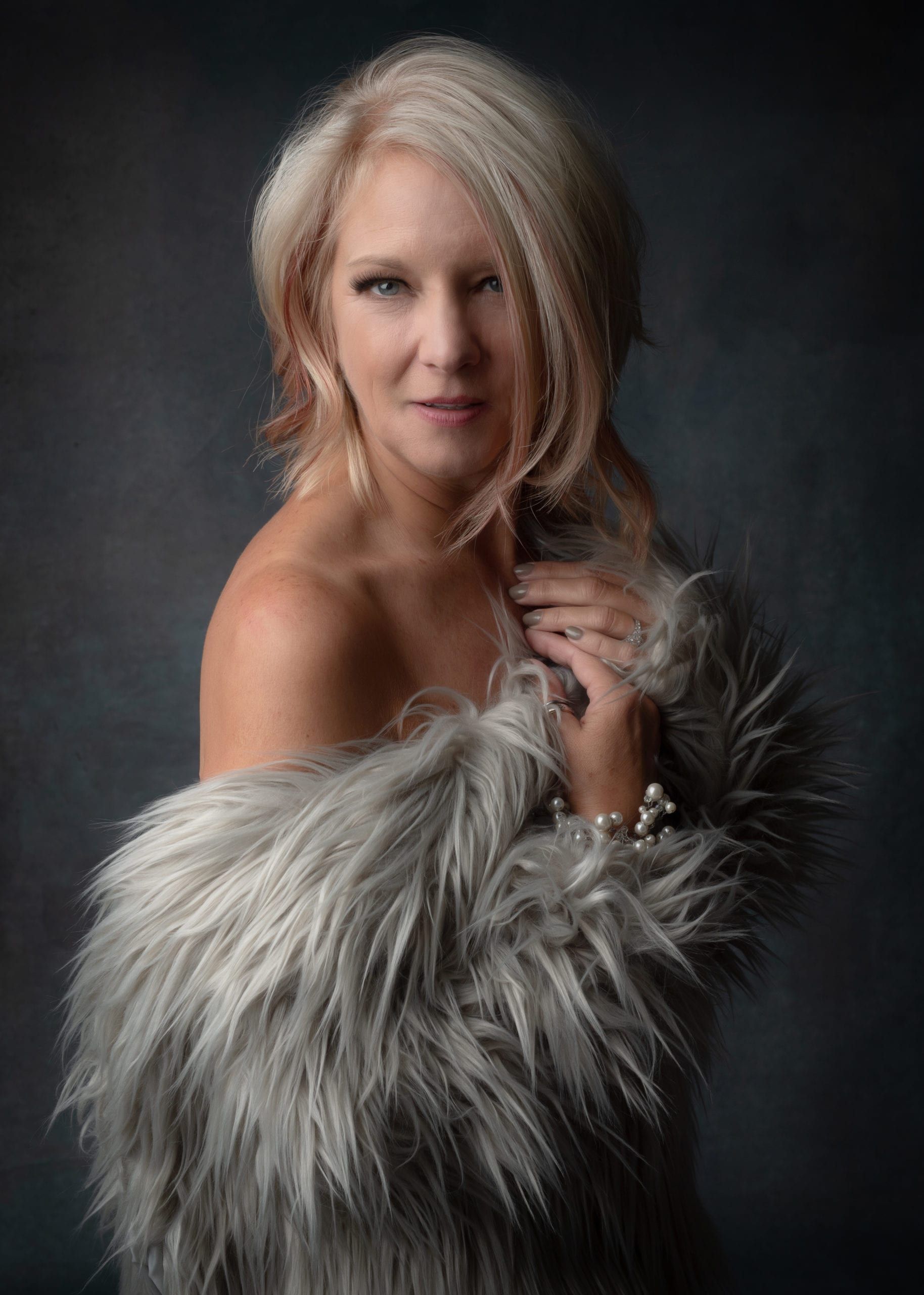 Blonde woman draped in a gray faux fur wrap, looking at the camera, against a dark gray backdrop.