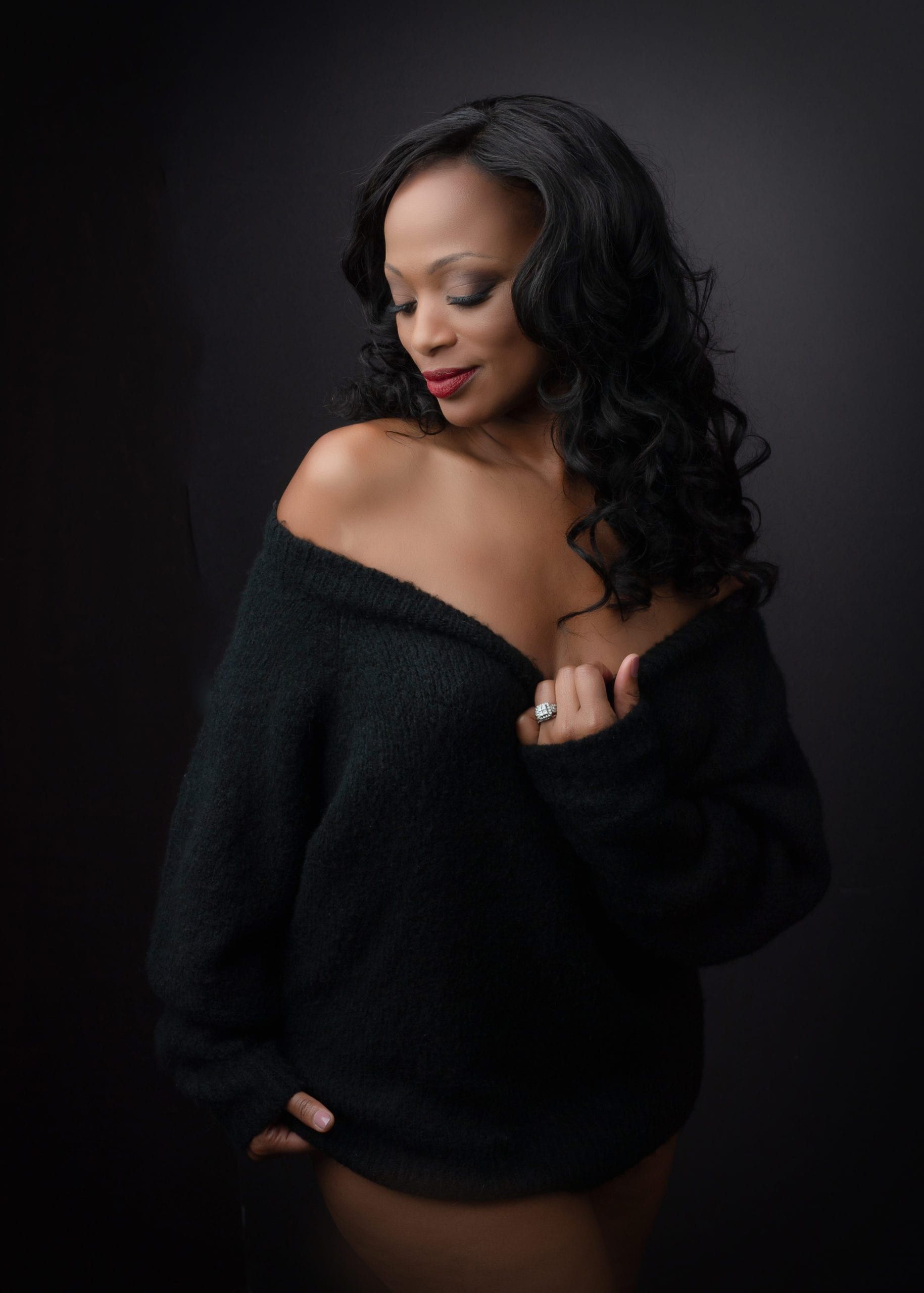 Woman in dark sweater, bare shoulders, looking down with dark hair and red lipstick.