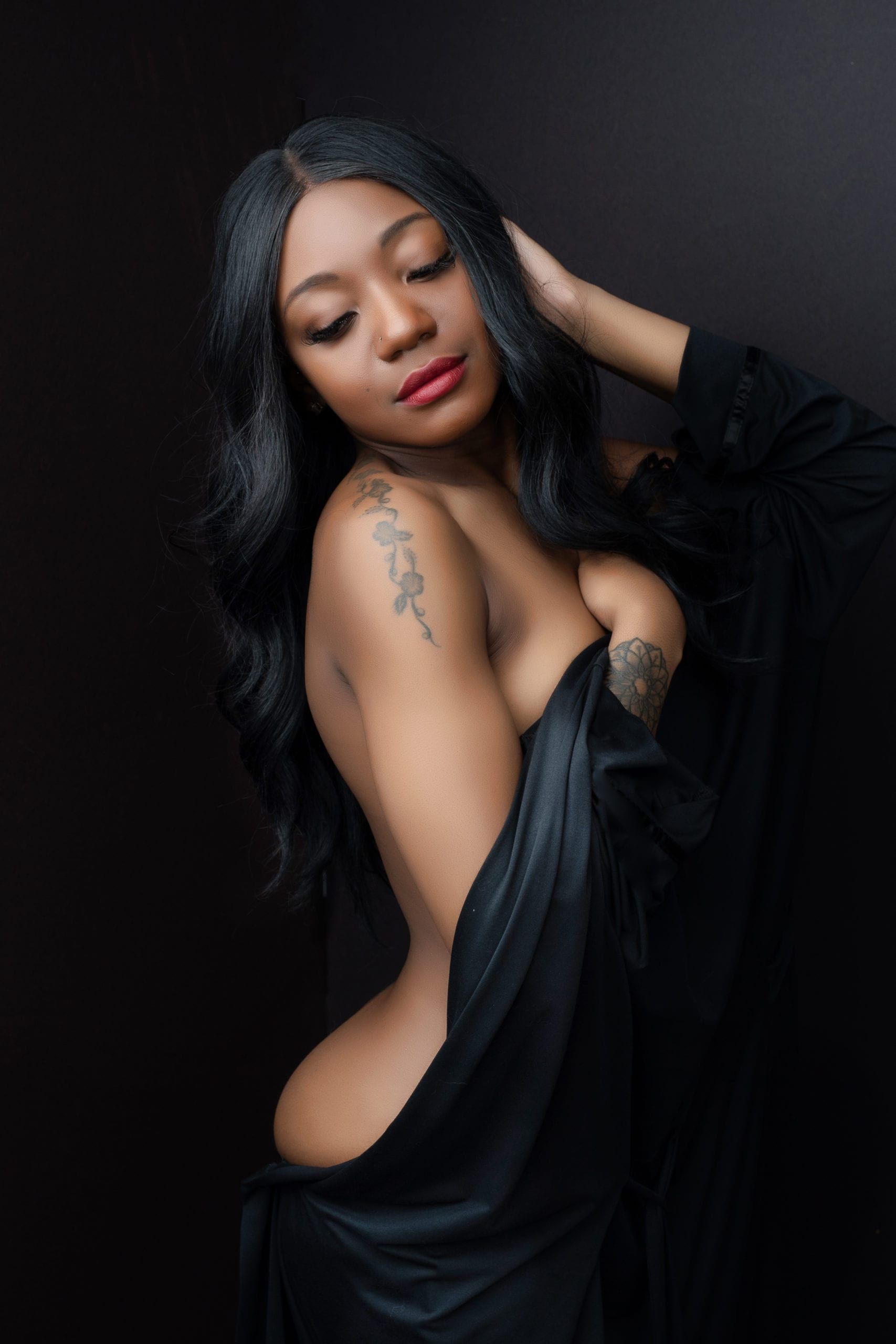Woman in black robe, partially nude. She has long black hair, a tattoo on her shoulder, and is looking down.