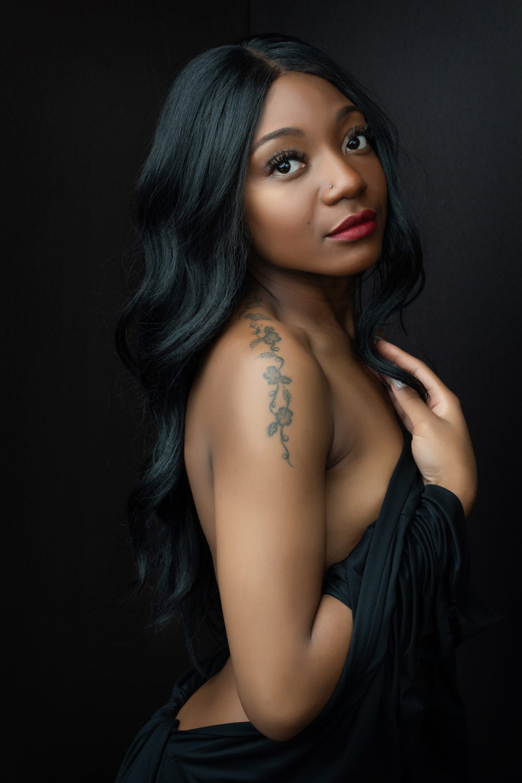 Black woman with long hair, looking at the viewer, holding black fabric, shoulder tattoo, dark background.