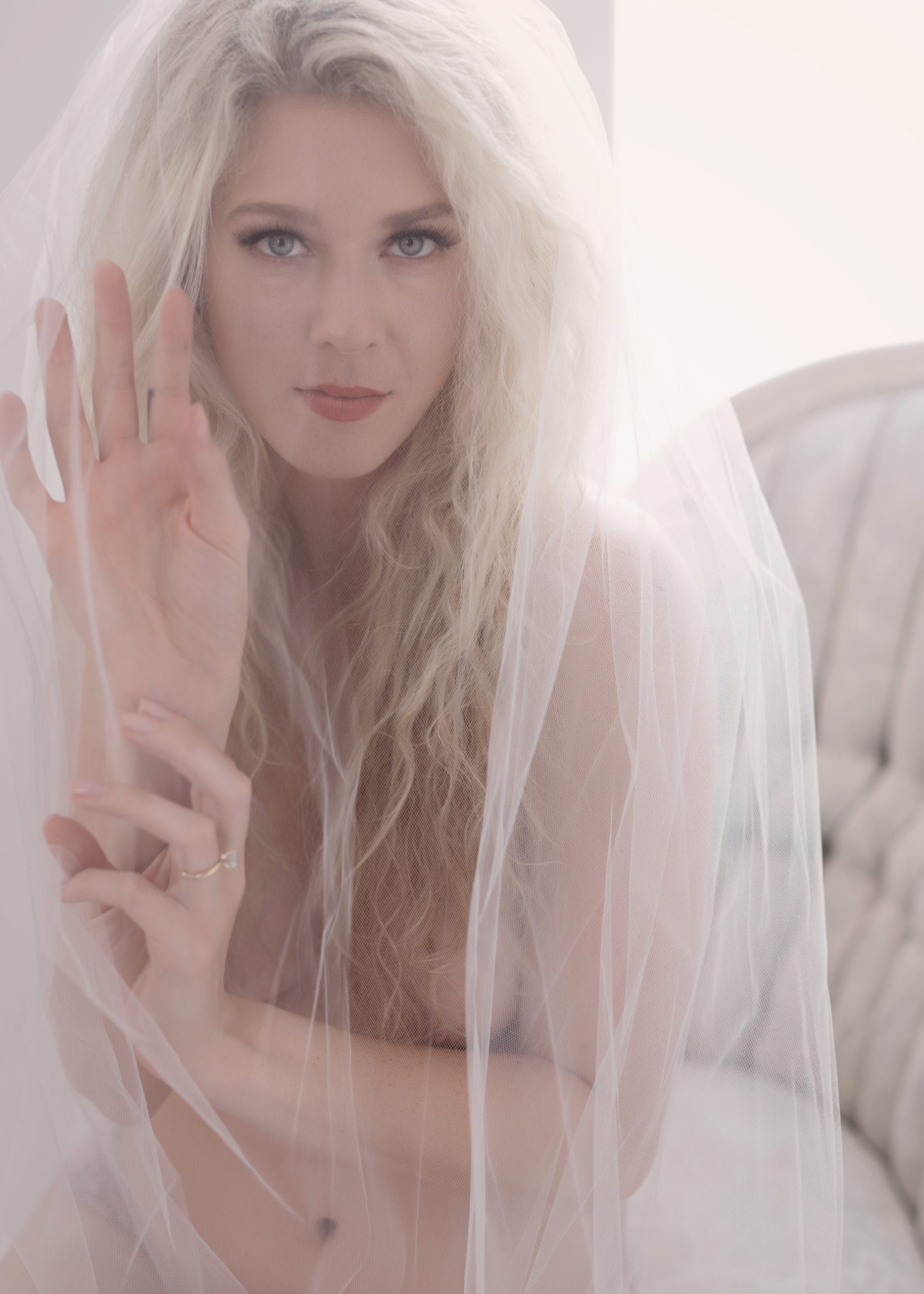 Blonde woman with curly hair veiled in white, hand raised, looking at the camera.