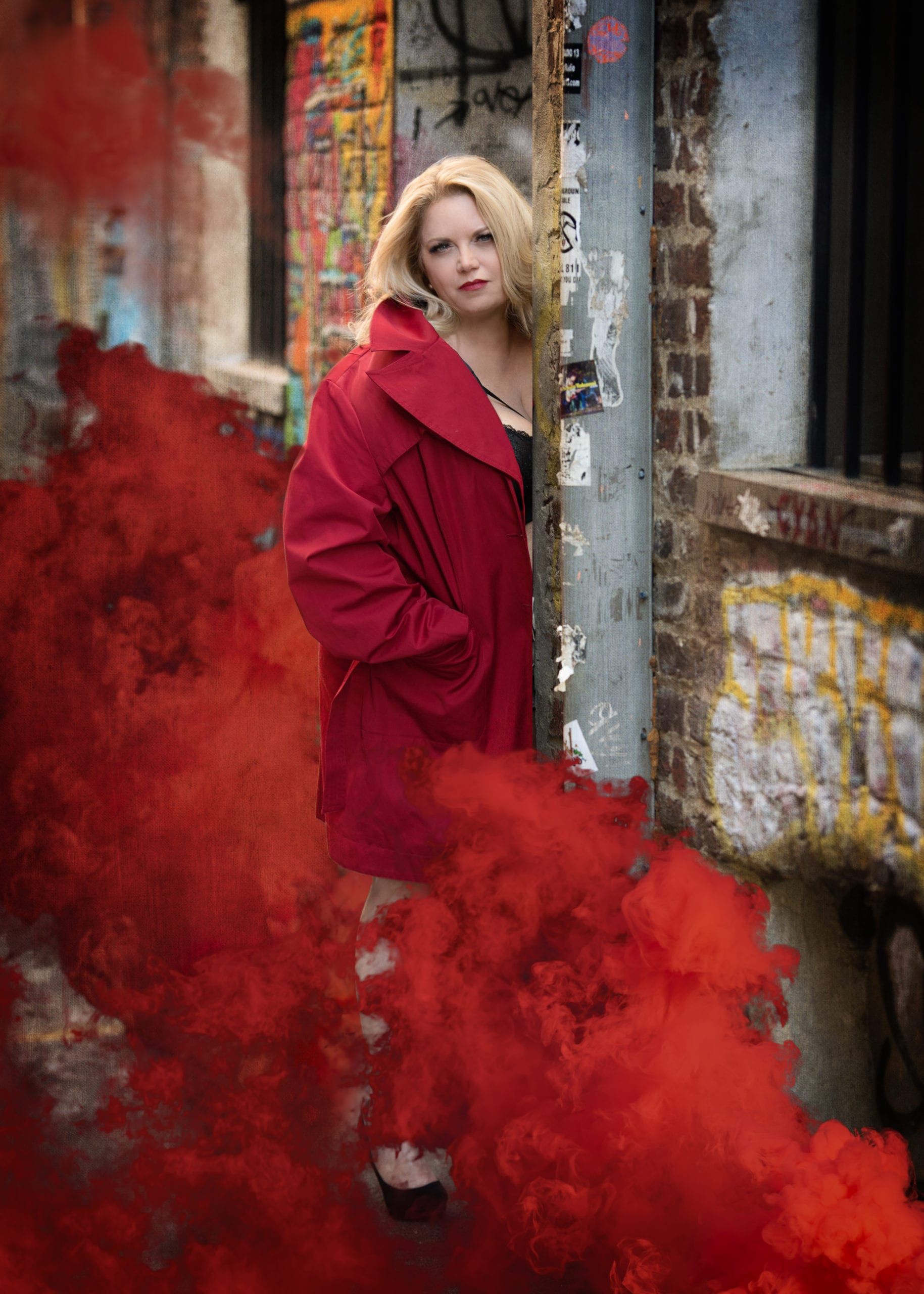 Blonde woman in red coat, red smoke around her, standing near a graffiti-covered wall.