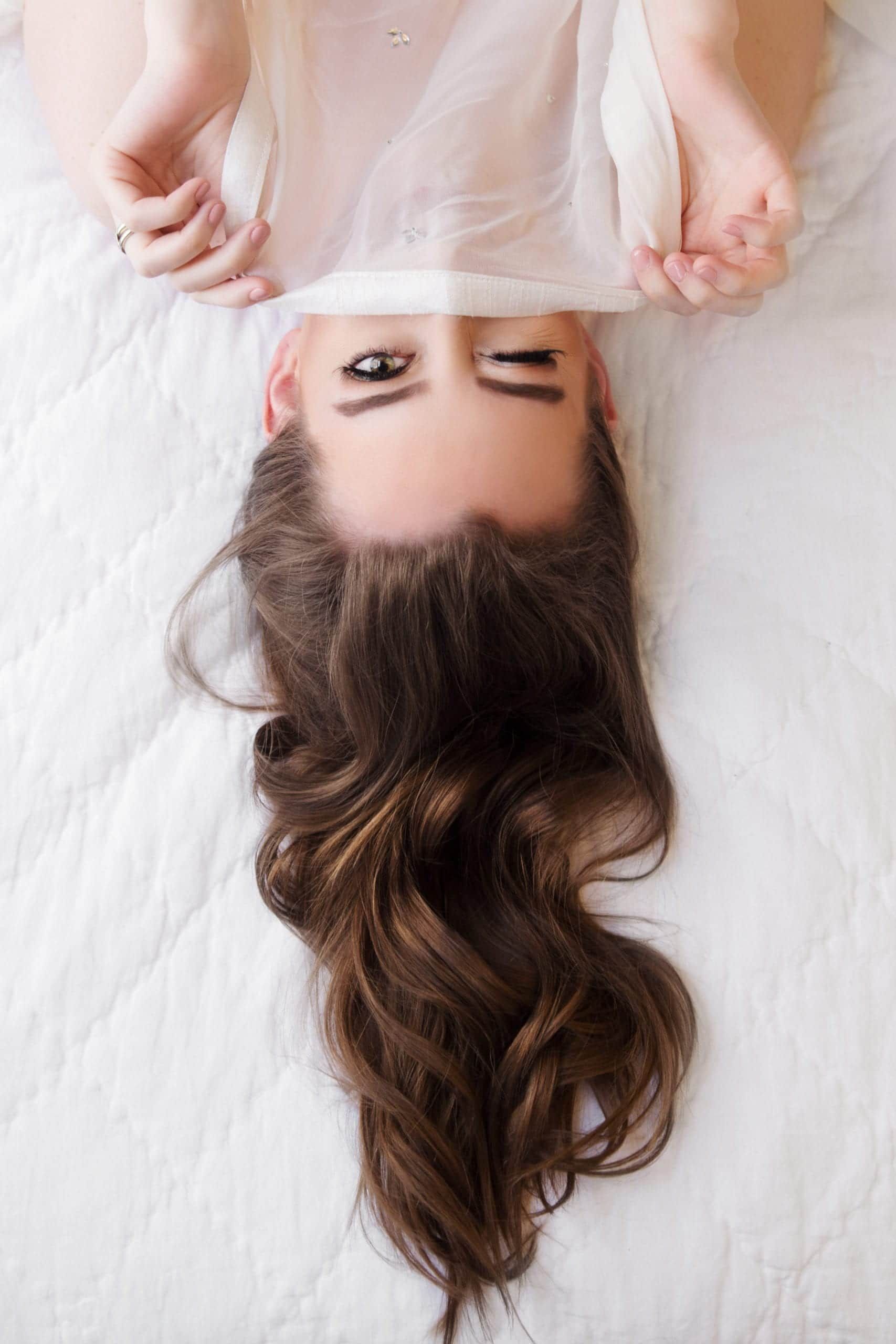 Woman with long brown hair, upside down on a white bed, holding up sheer fabric to partially cover her face.