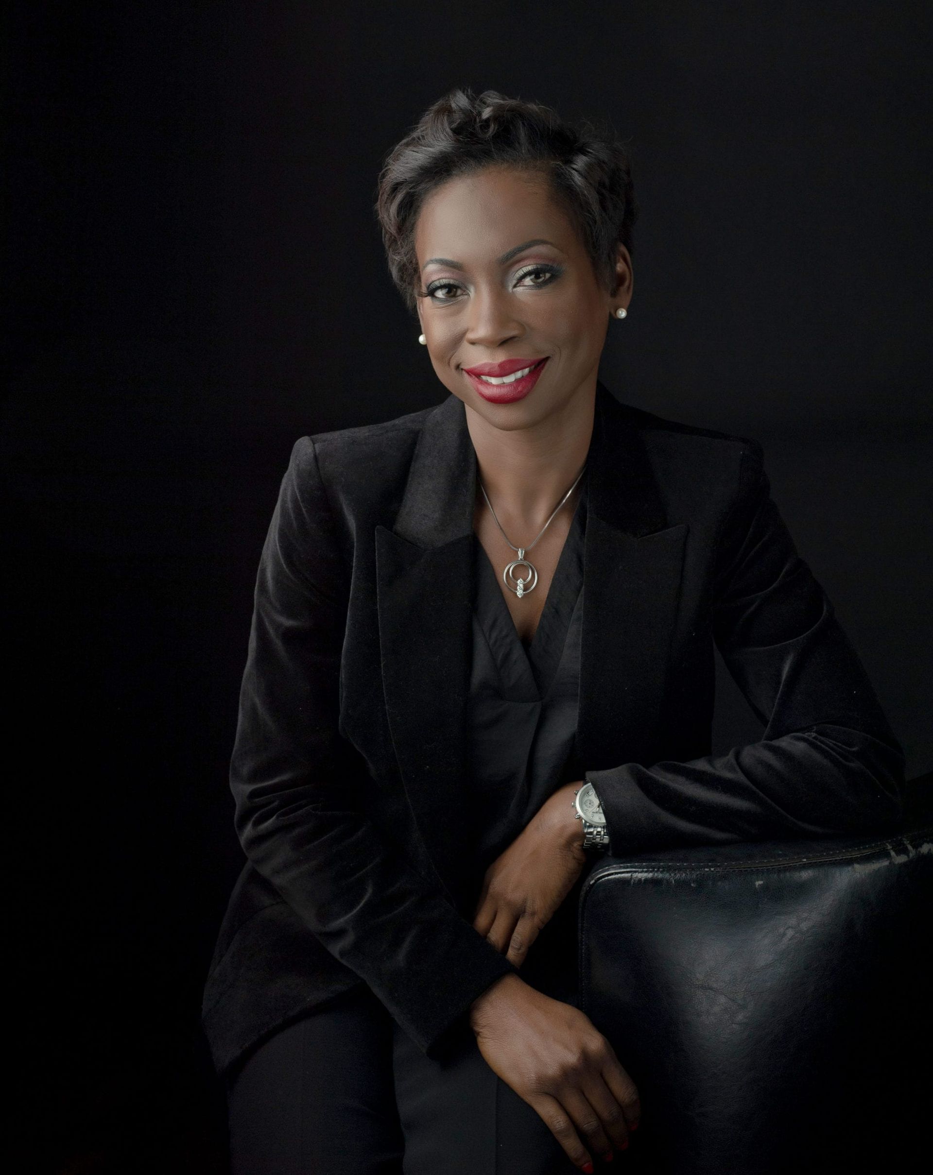 Woman in black suit, smiling, resting arm on chair. Black background.