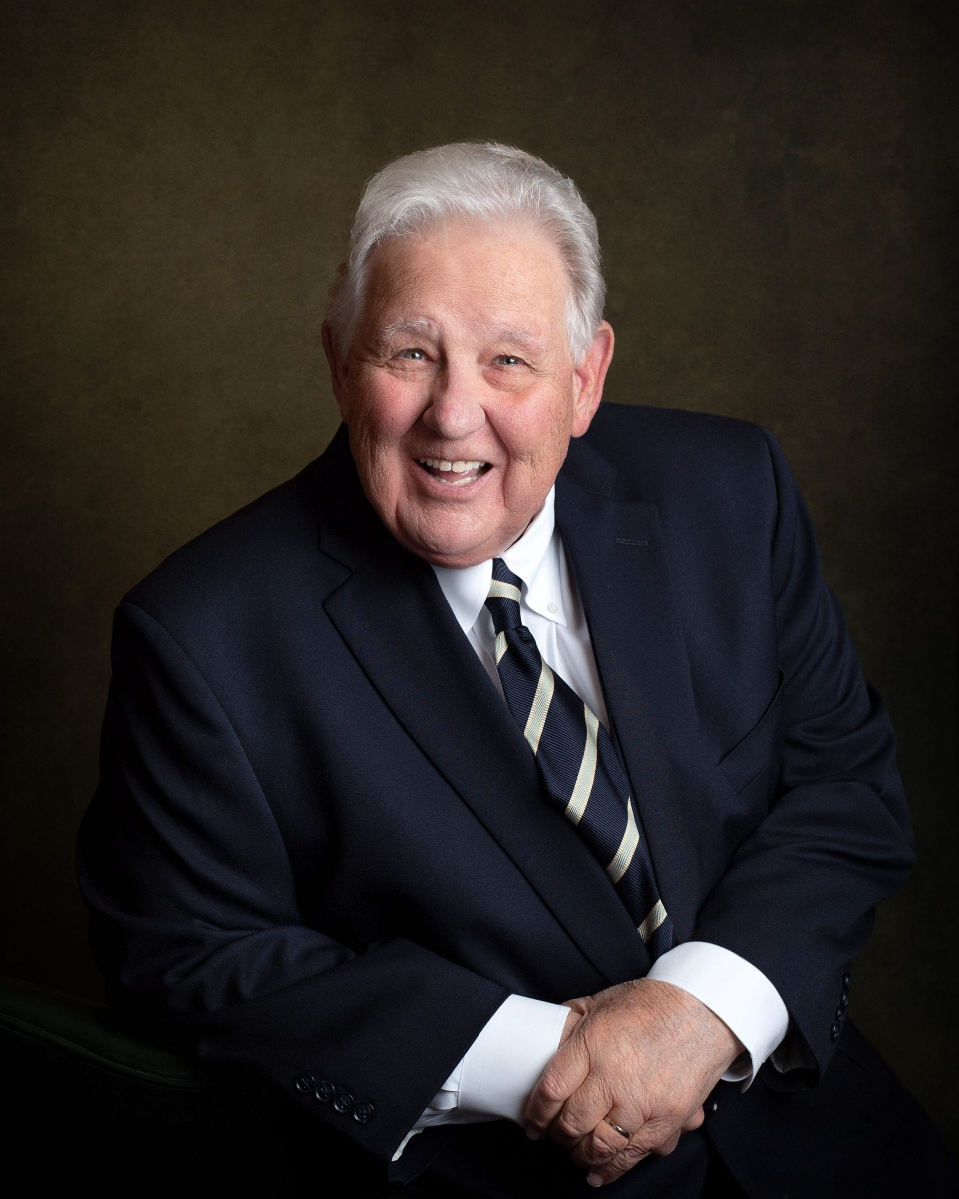 Older man with white hair smiles, wearing a navy suit and striped tie, in a studio setting.