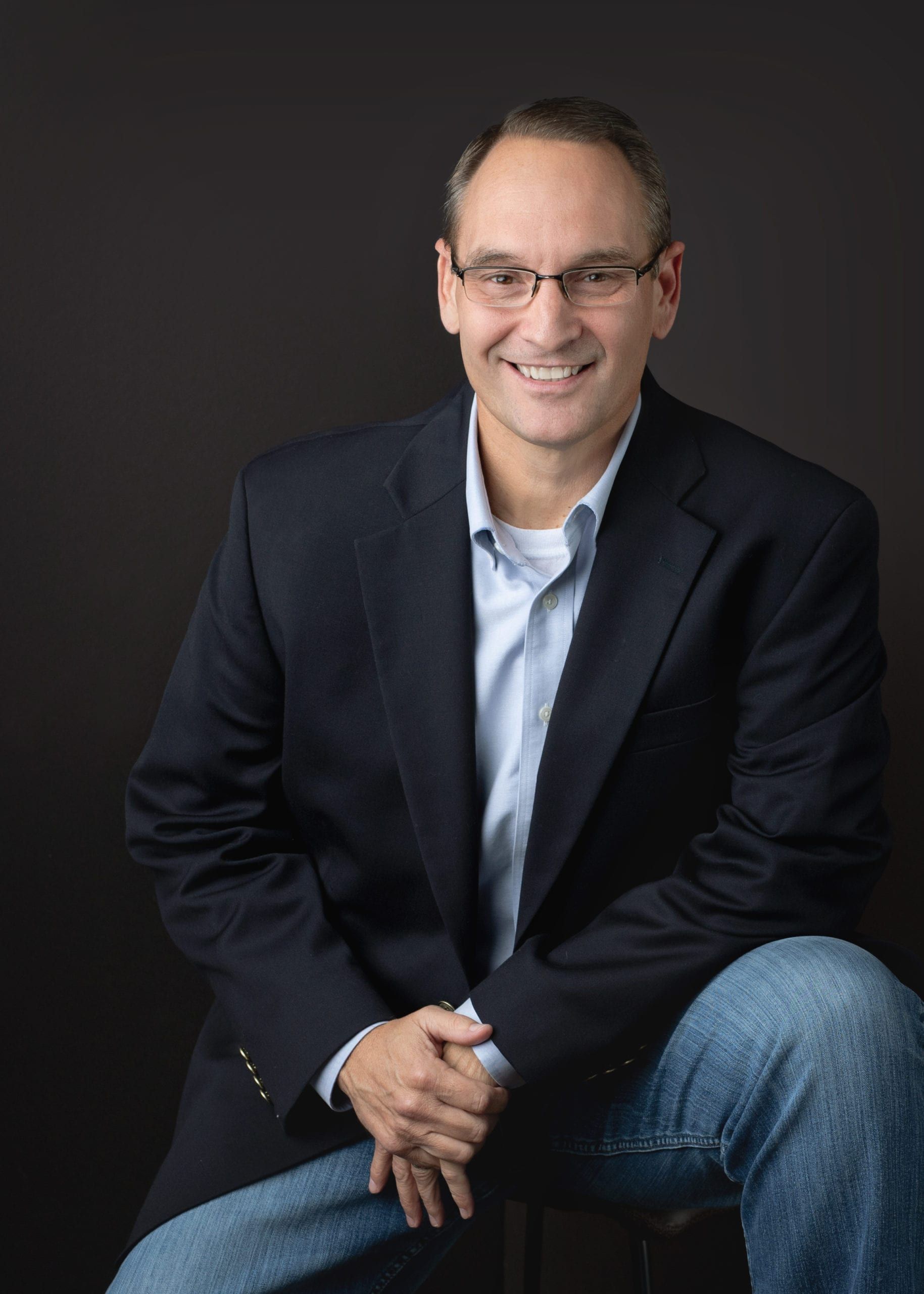 Man in blazer and jeans smiles while leaning, against a black background.
