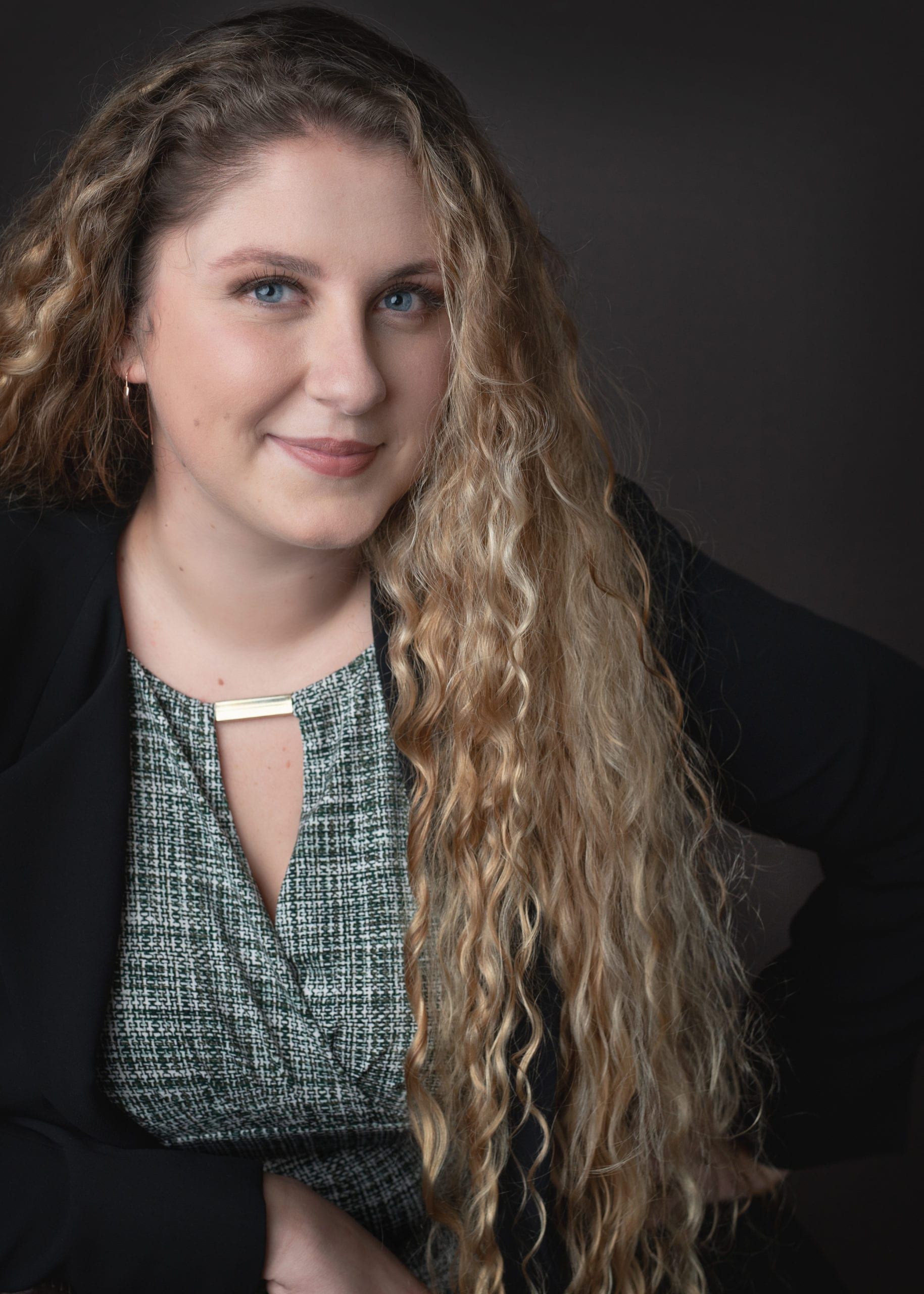 Woman with long, curly blonde hair, smiling, wearing a green and black top and black blazer.