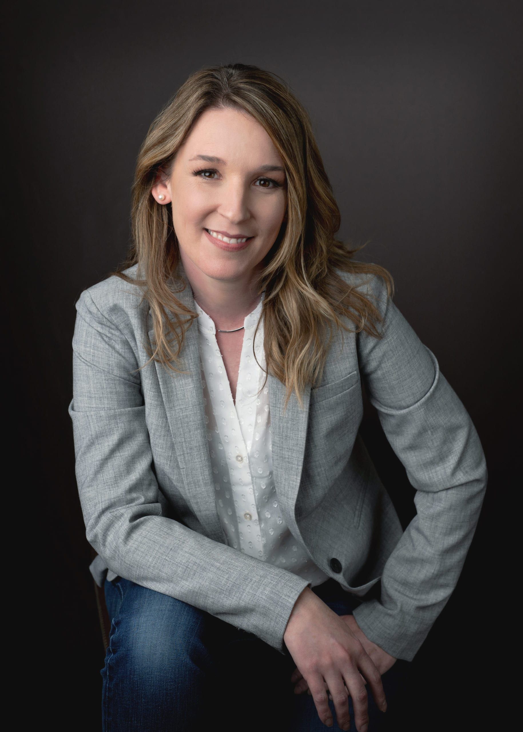 Woman in a gray blazer and jeans smiles against a dark background, sitting and leaning forward.