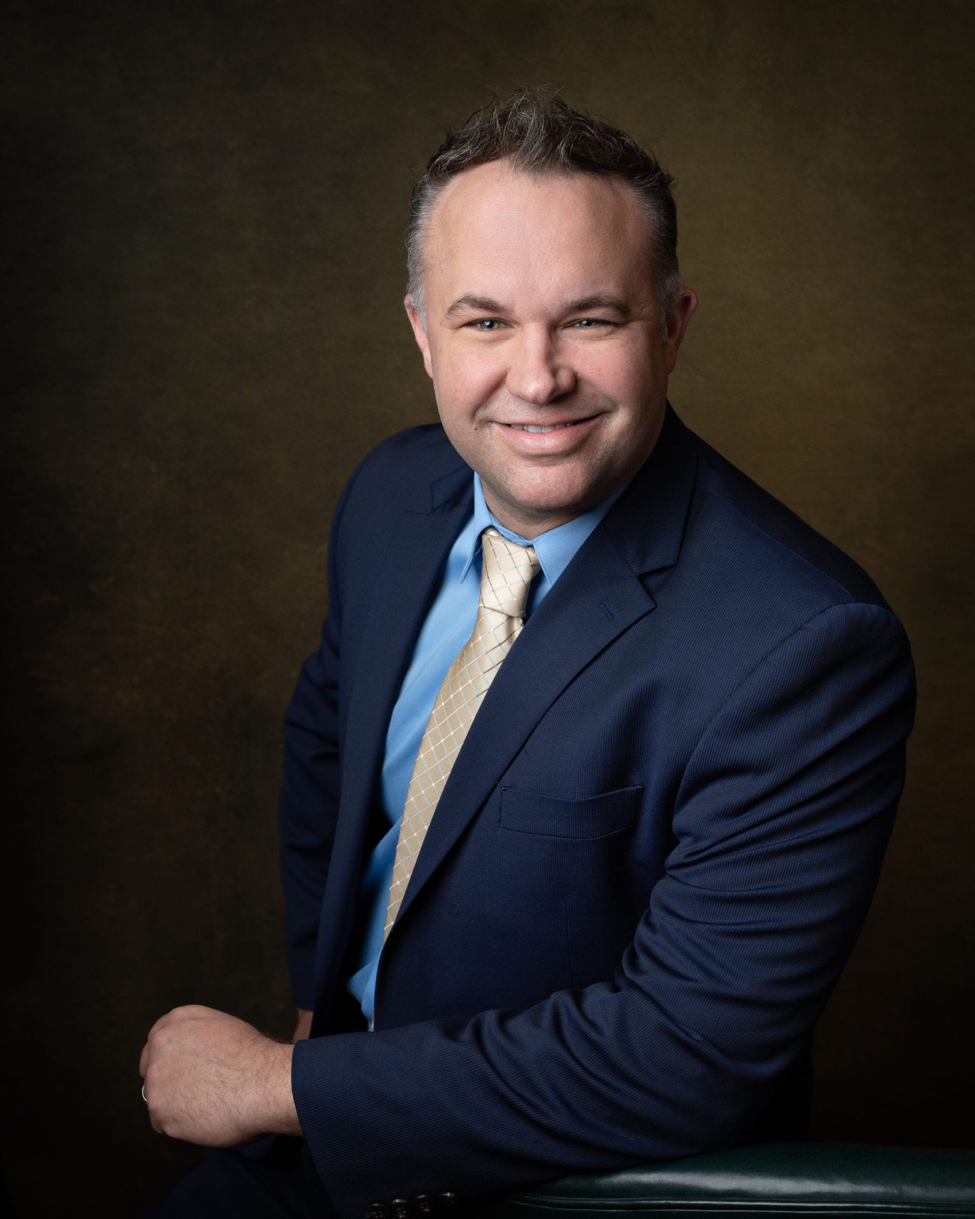 Man in blue suit and tie smiles, leaning on a dark background.