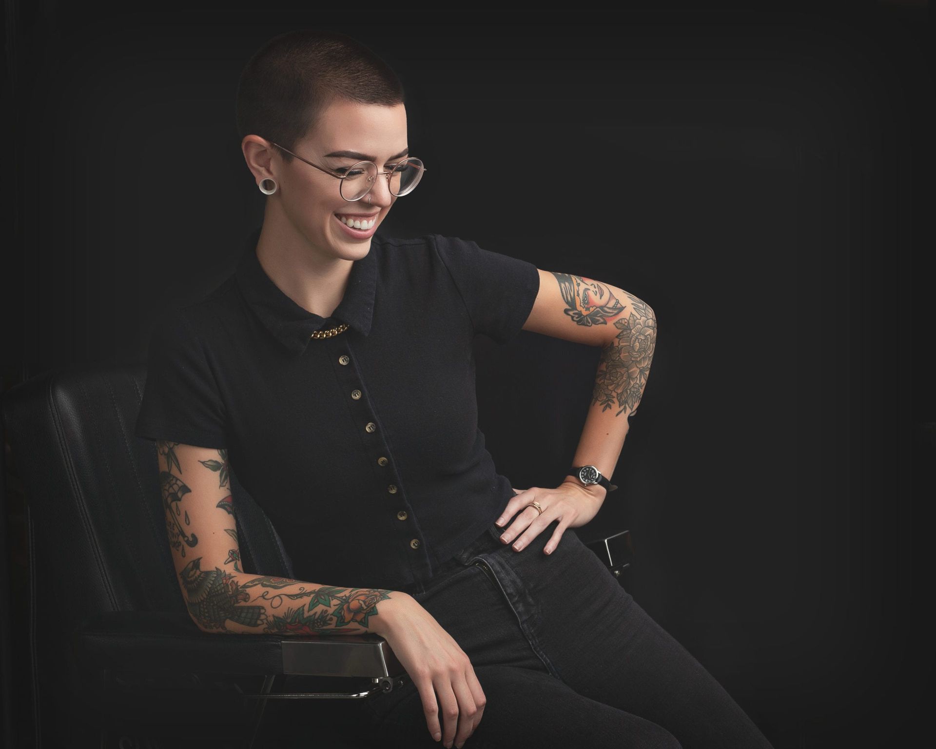 Woman with shaved head, tattoos, and glasses, smiling, seated, wearing black clothes, against a black background.