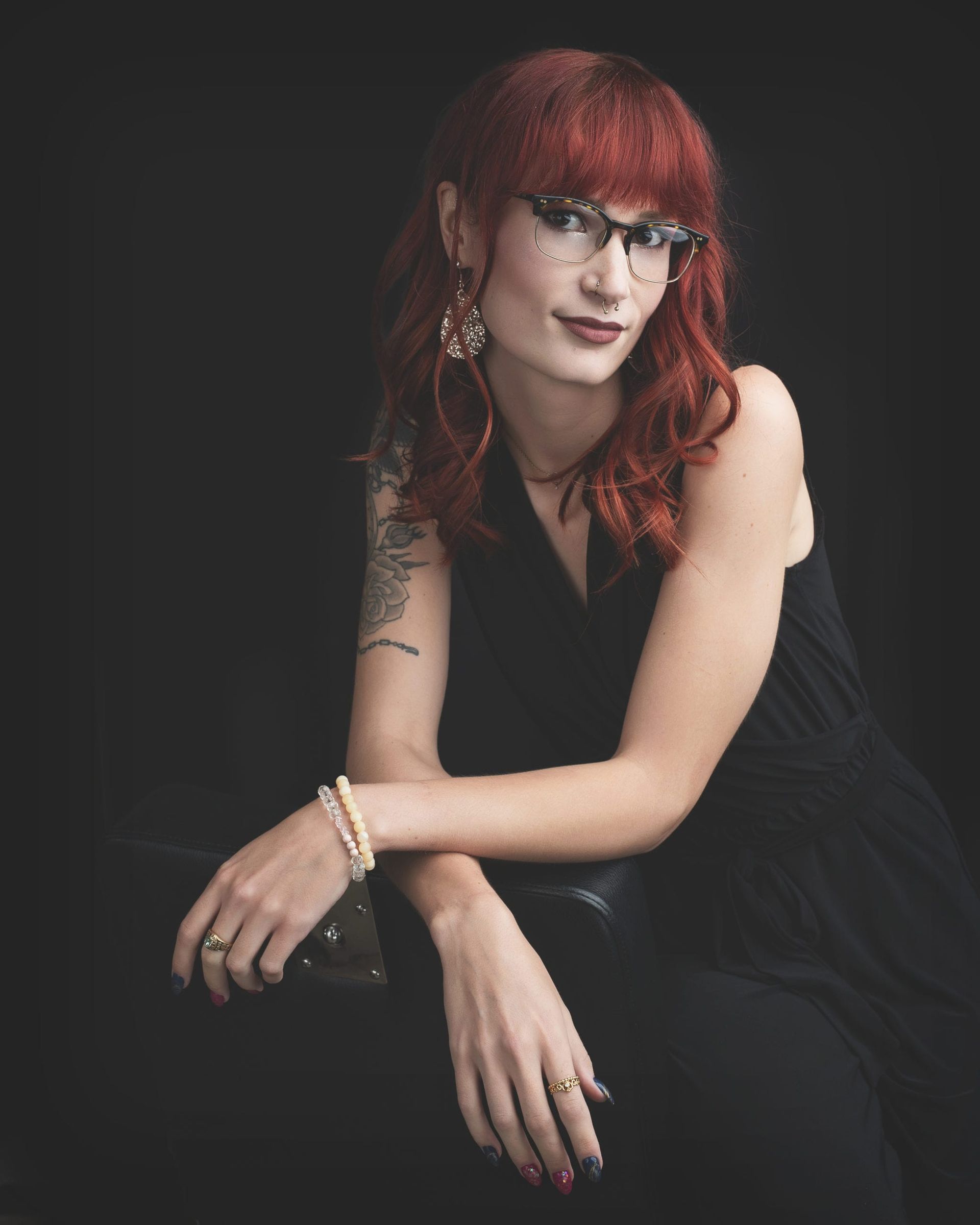 Woman with red hair and glasses leans, wearing black dress, dark background.