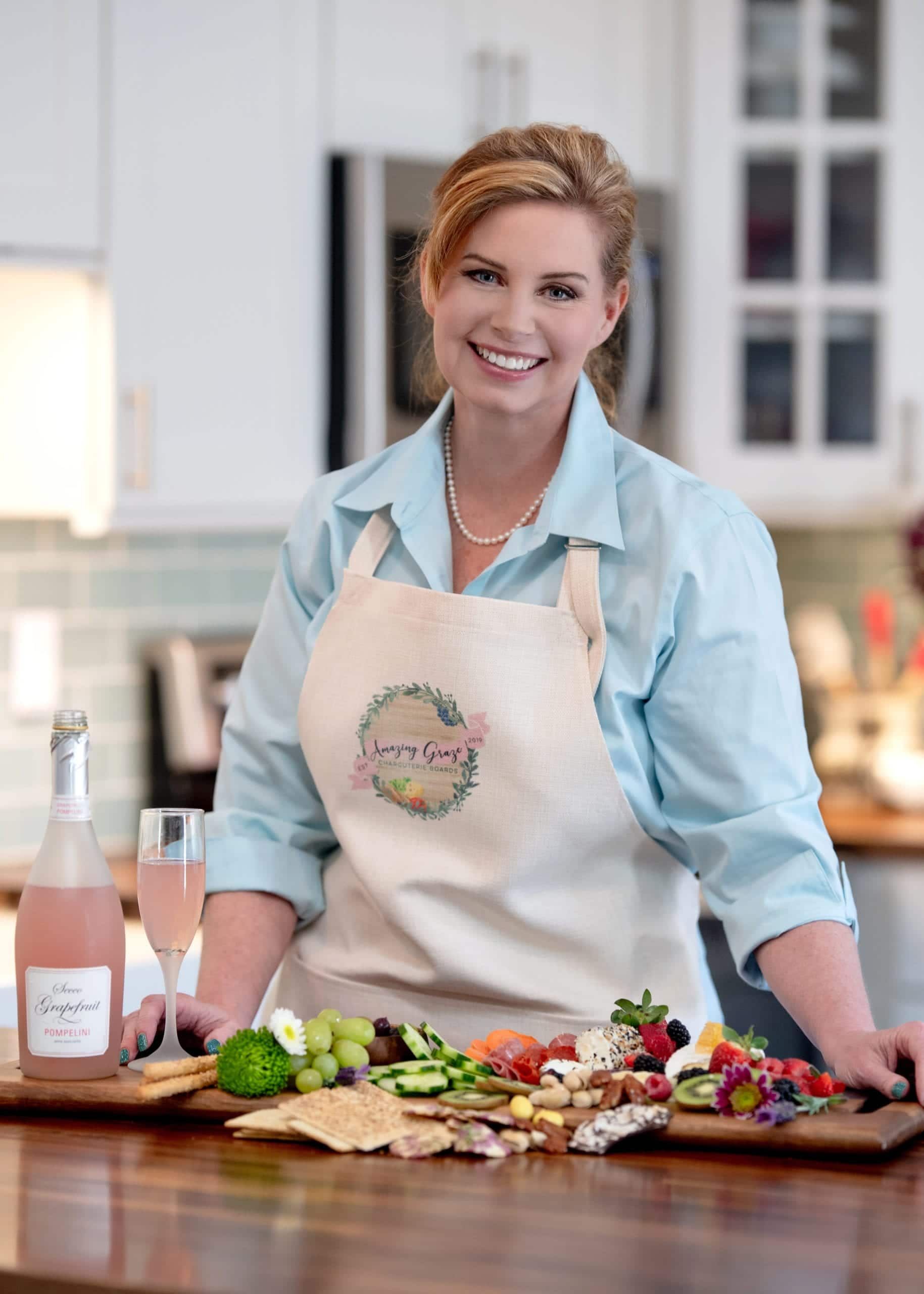 Woman in apron smiles at the camera, beside a charcuterie board and pink champagne in a kitchen.