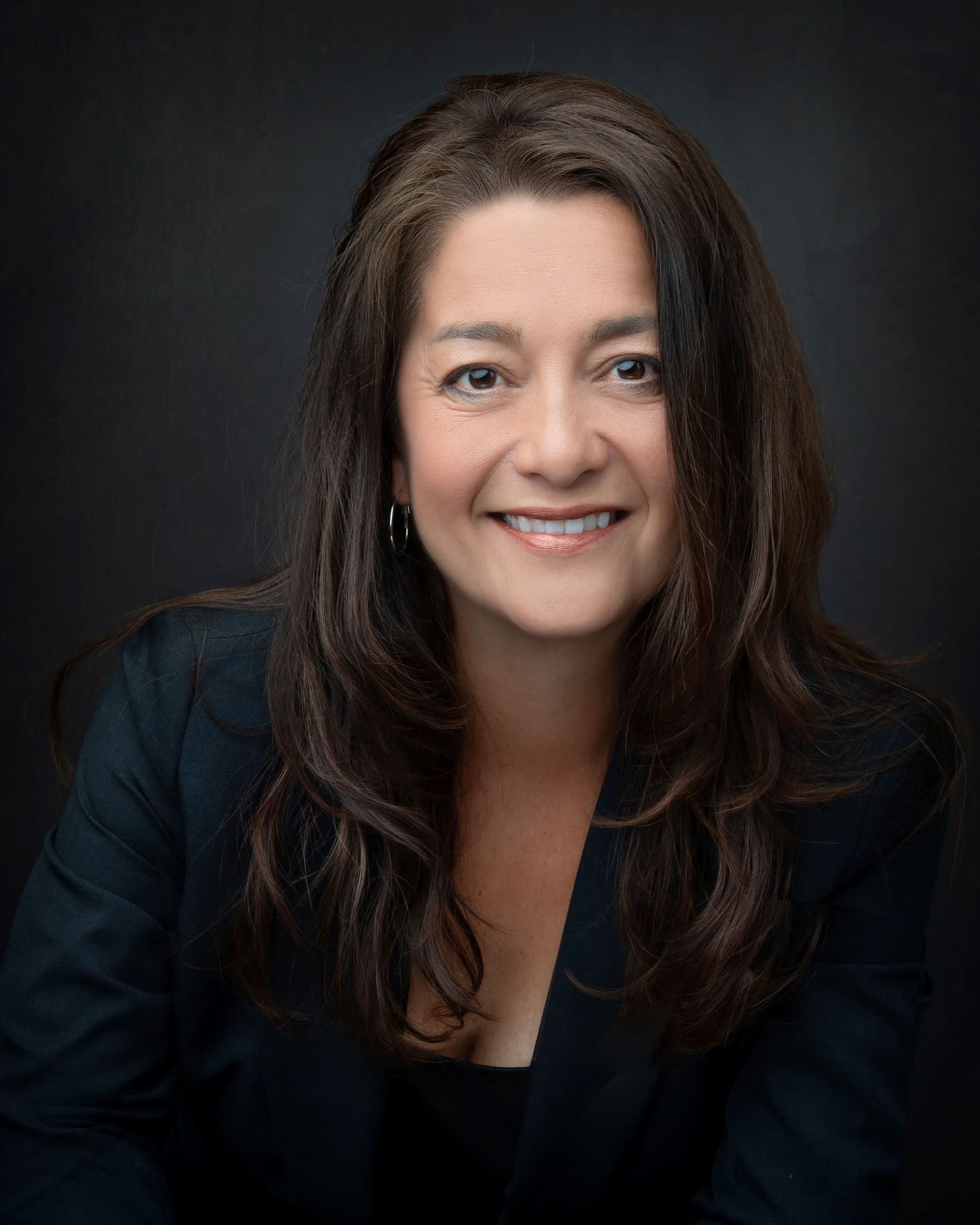Woman with long dark hair smiling, wearing a dark blazer against a dark background.