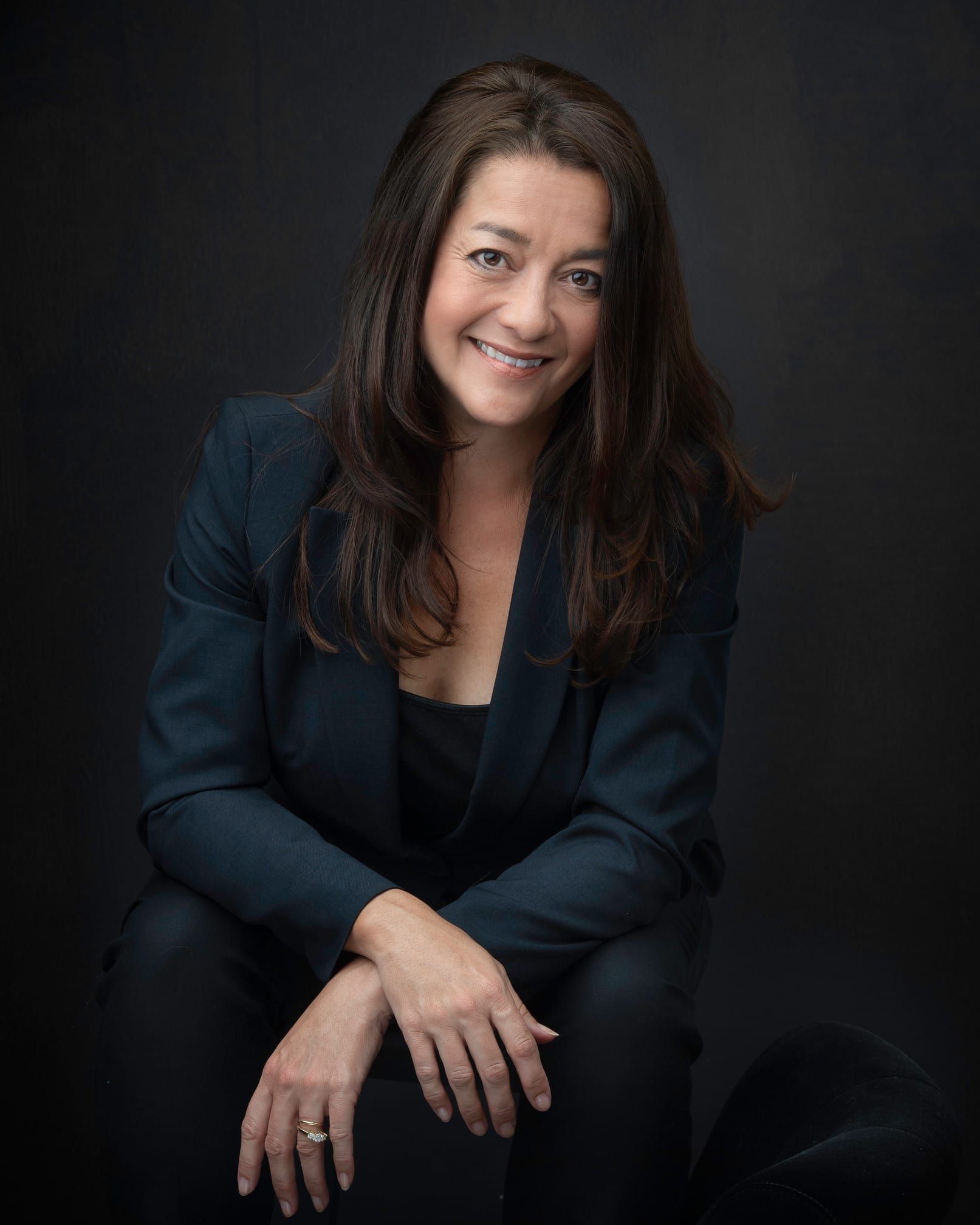 Woman in dark suit smiles, sitting with arms crossed; dark background.