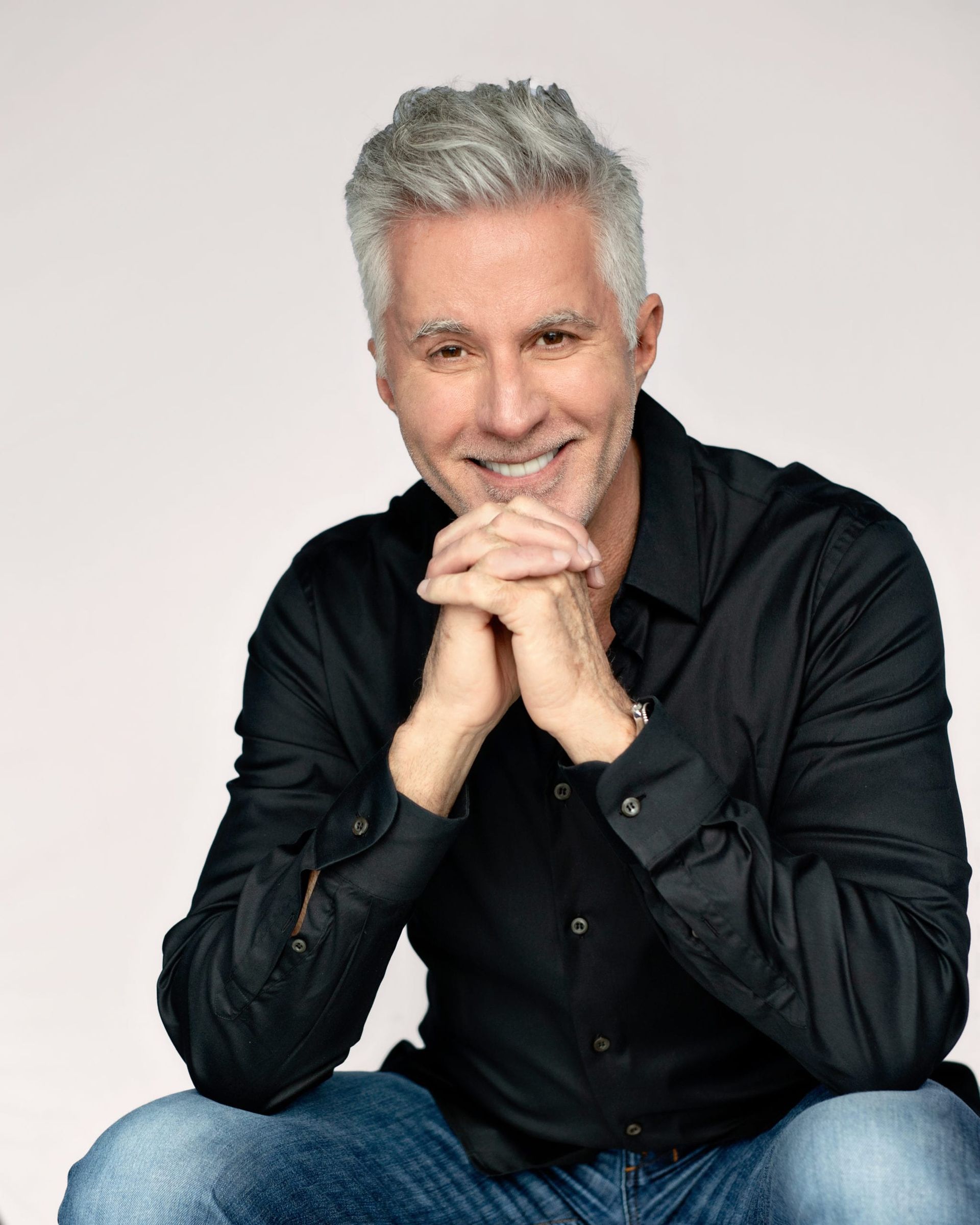 Man with gray hair, hands clasped, smiling, wearing a black shirt and blue jeans.