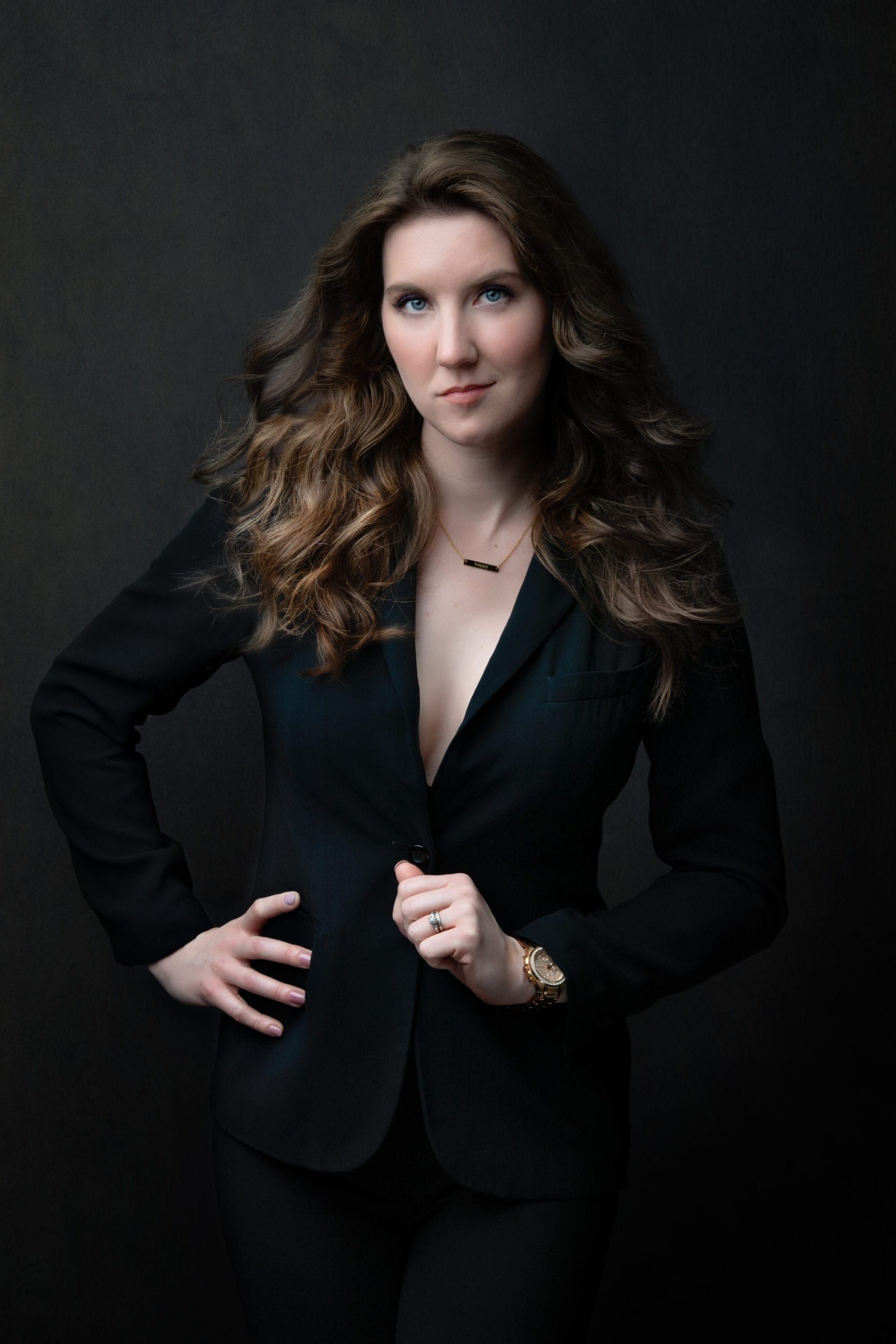Woman in black suit, hand on hip, looking at camera, dark background.