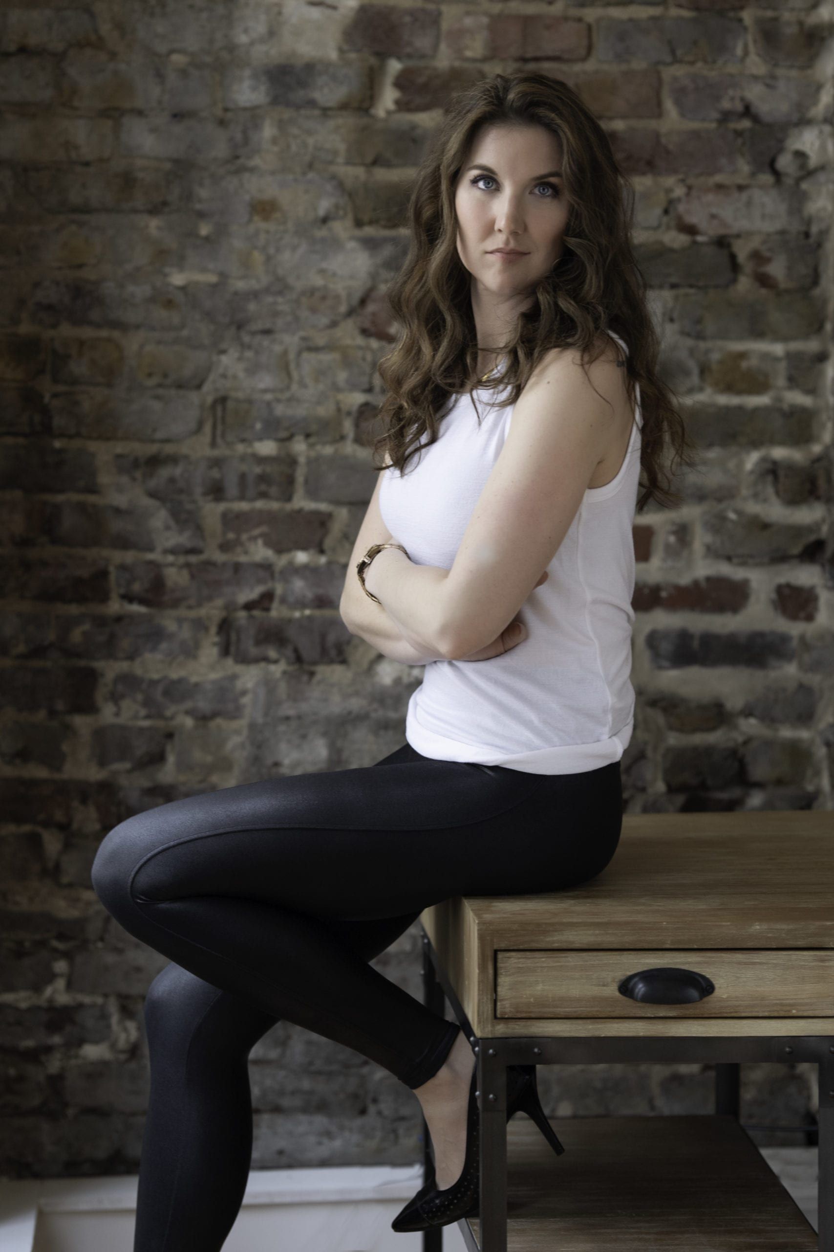 Woman in white top and black pants sitting on a table, arms crossed, against a brick wall.