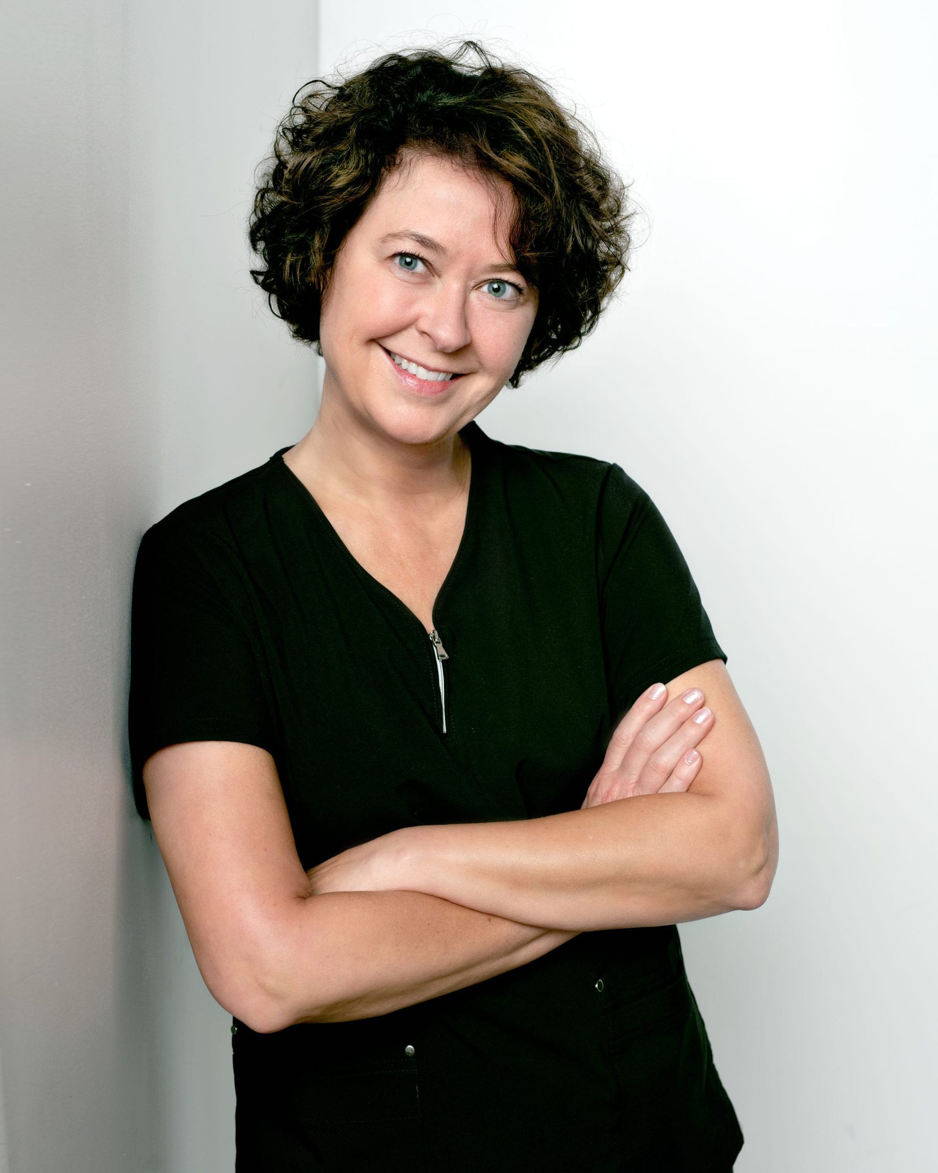 Woman with curly brown hair smiles, arms crossed. Leaning against a white wall, wearing a black shirt.