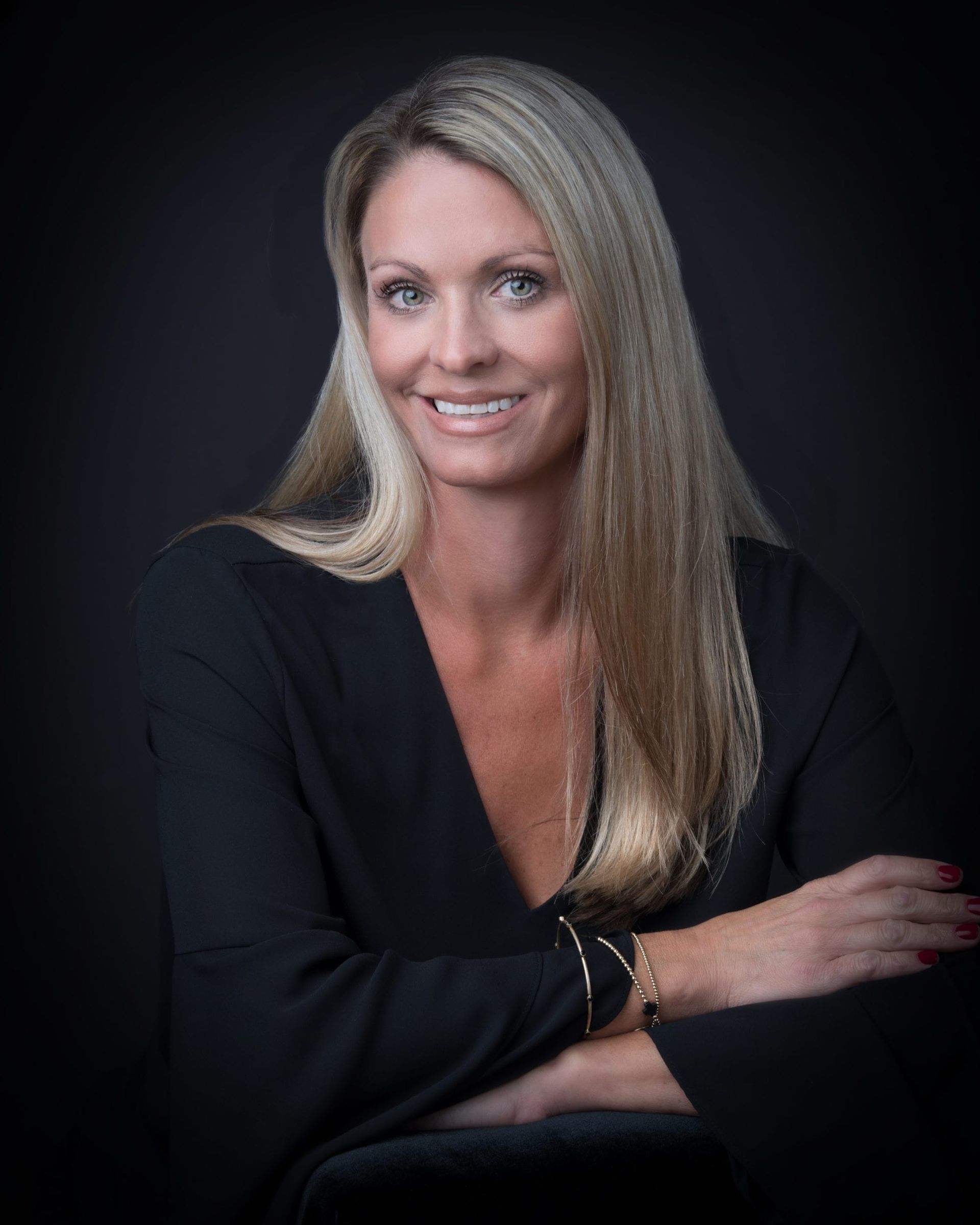 Blonde woman with arms crossed, smiles at camera, wearing black dress. Black background.