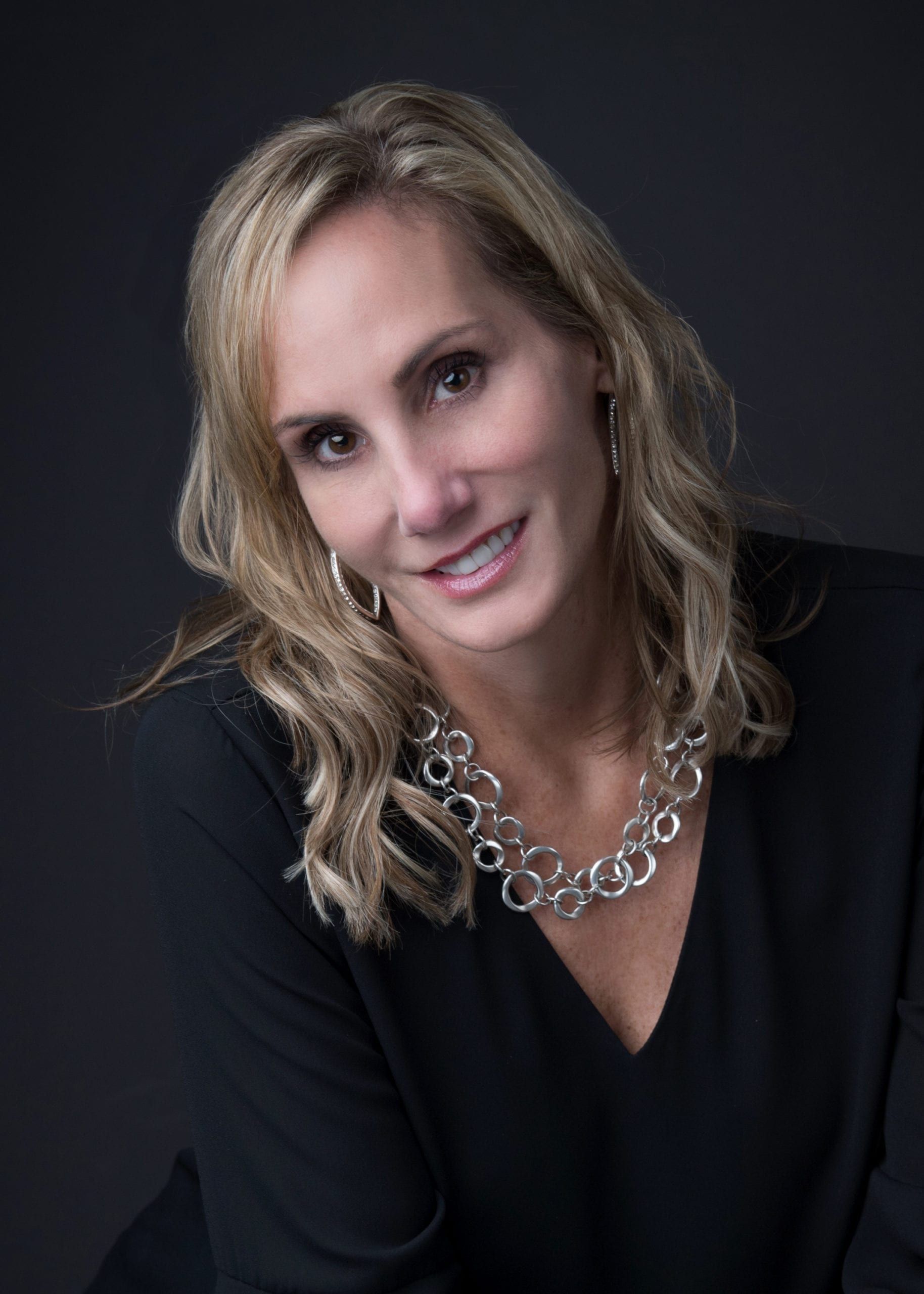 Blonde woman smiles at the camera. She wears a black top and a silver necklace, against a dark backdrop.