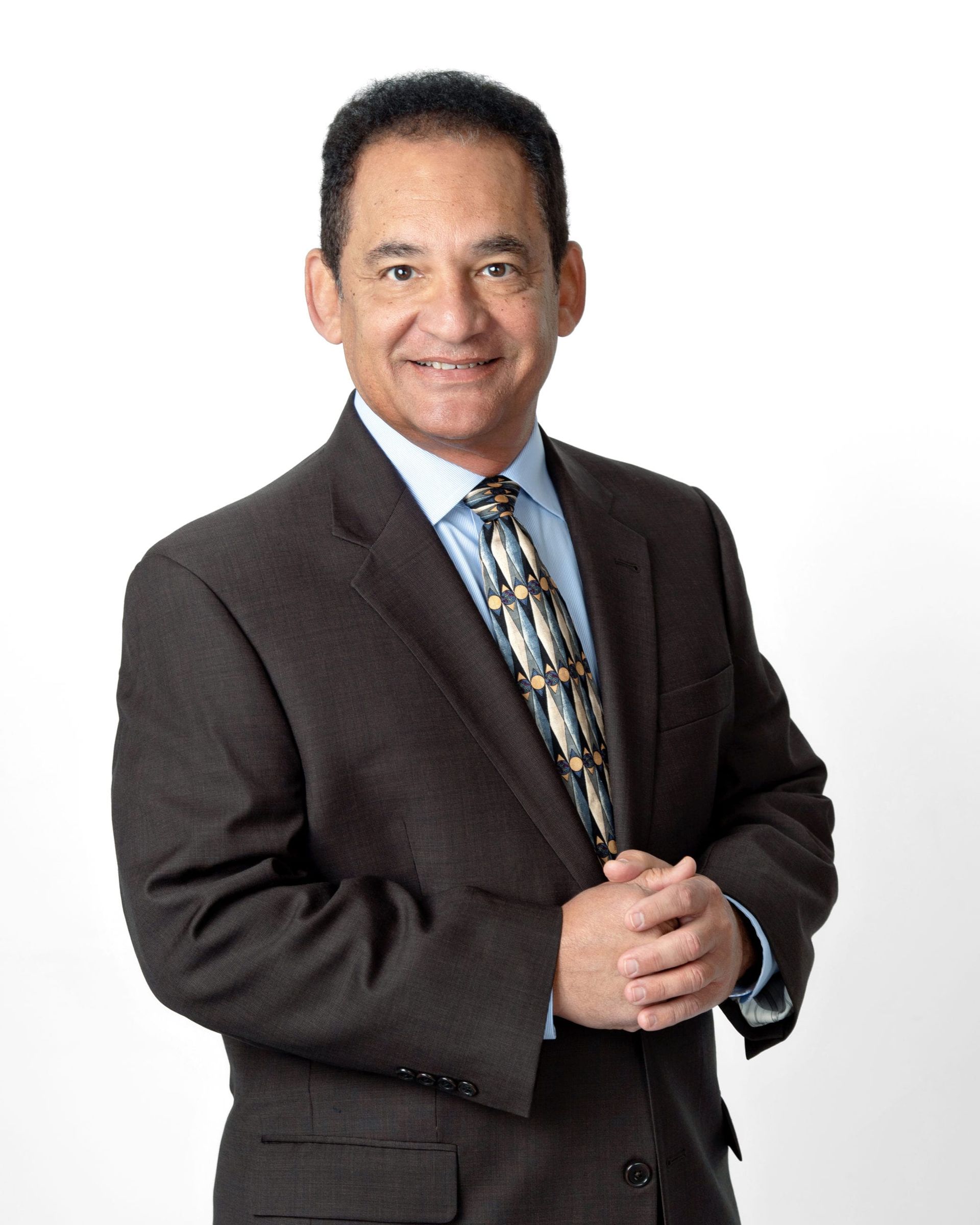 Man in suit, smiling, hands clasped, neutral background.