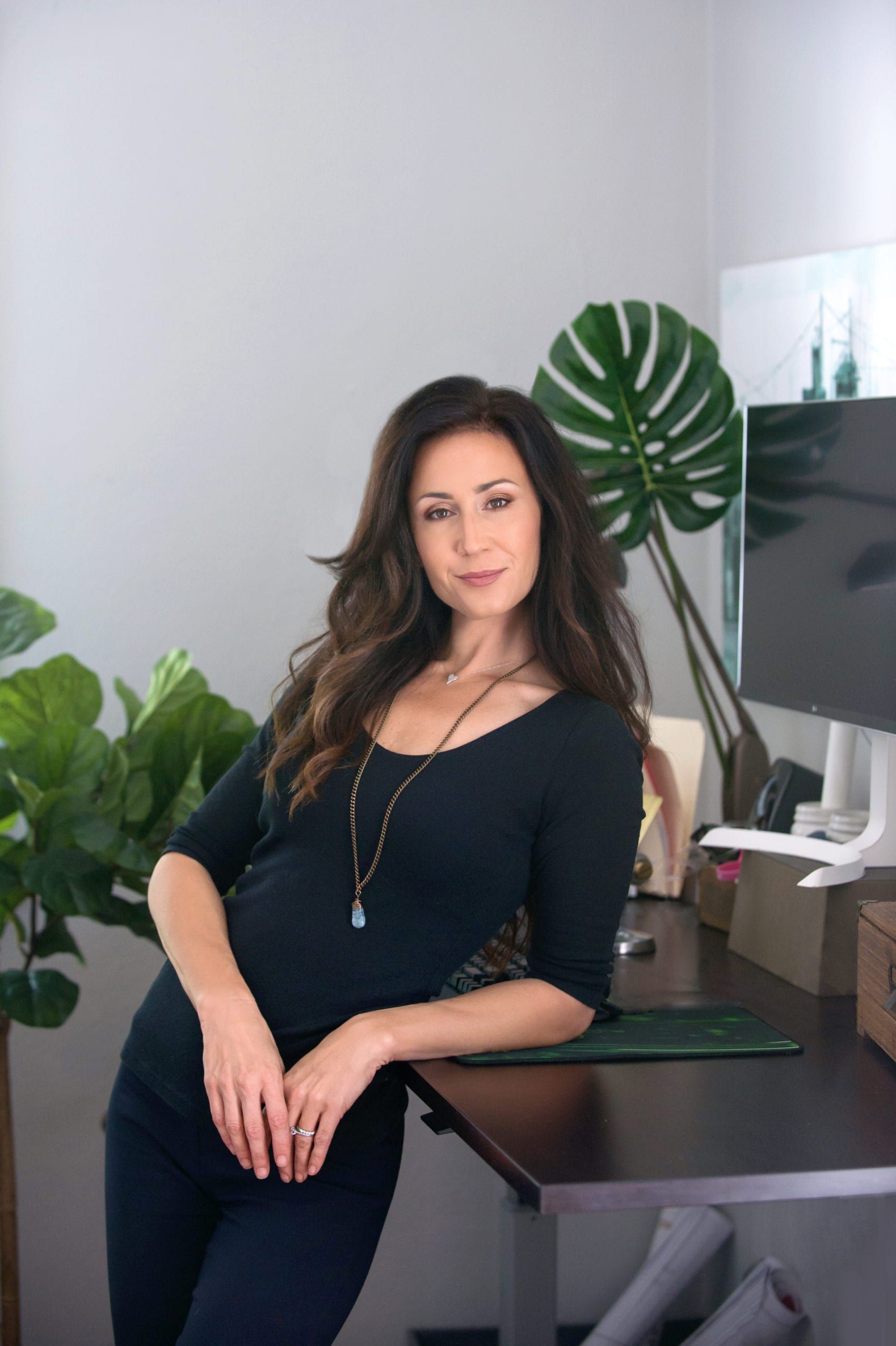 Woman with long brown hair leans on a desk in an office, smiling.