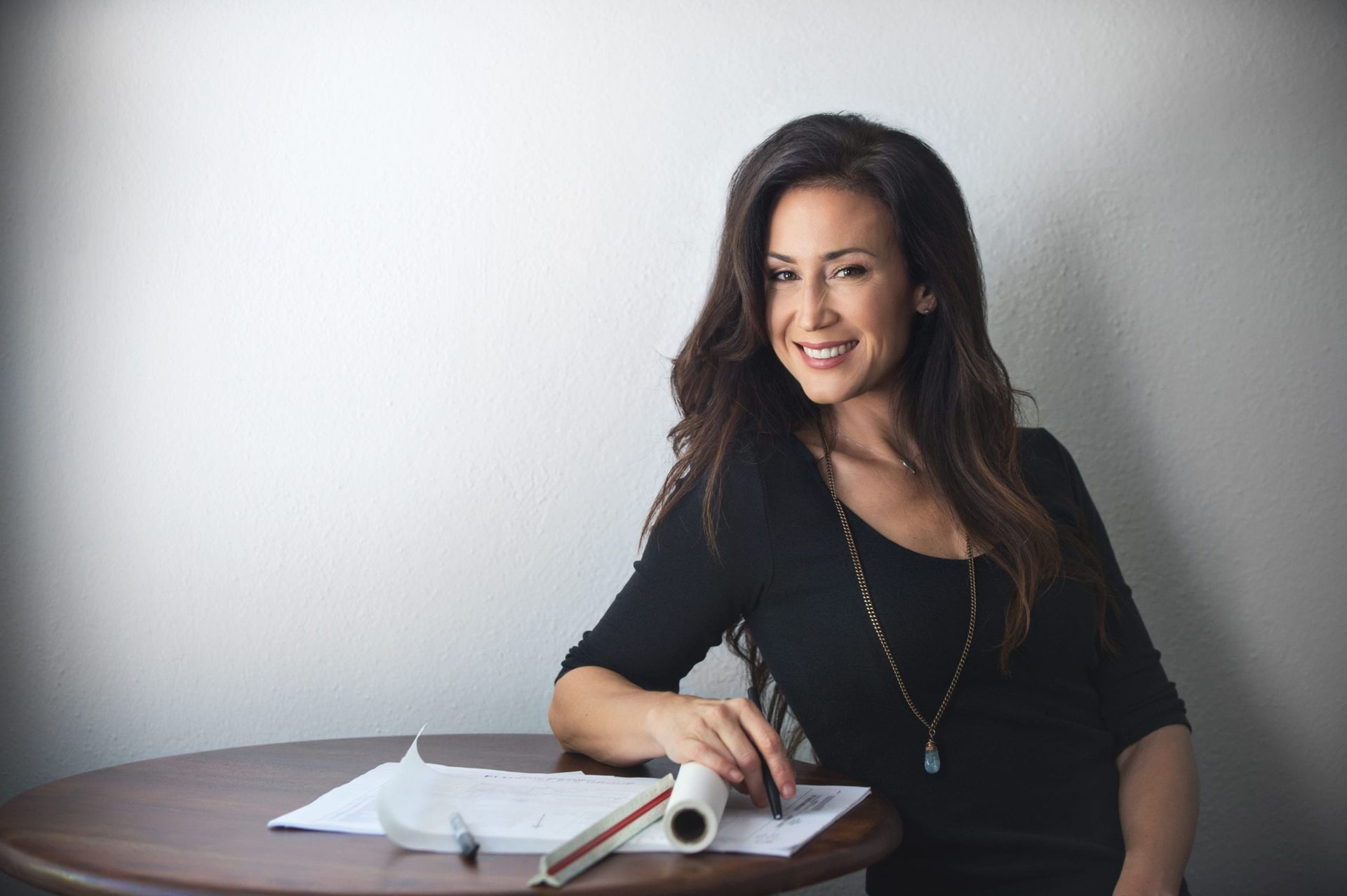 Woman with long brown hair smiling, sitting at a table with papers and a pen, leaning against a white wall.