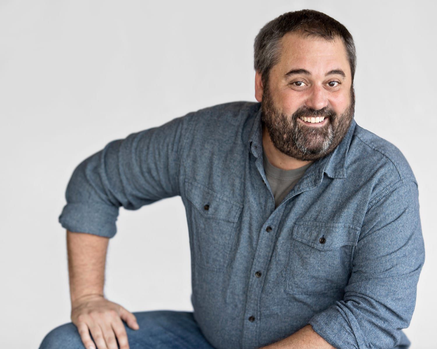 Man with a beard, smiling, wearing a blue shirt, sitting with one knee raised. Against a white background.
