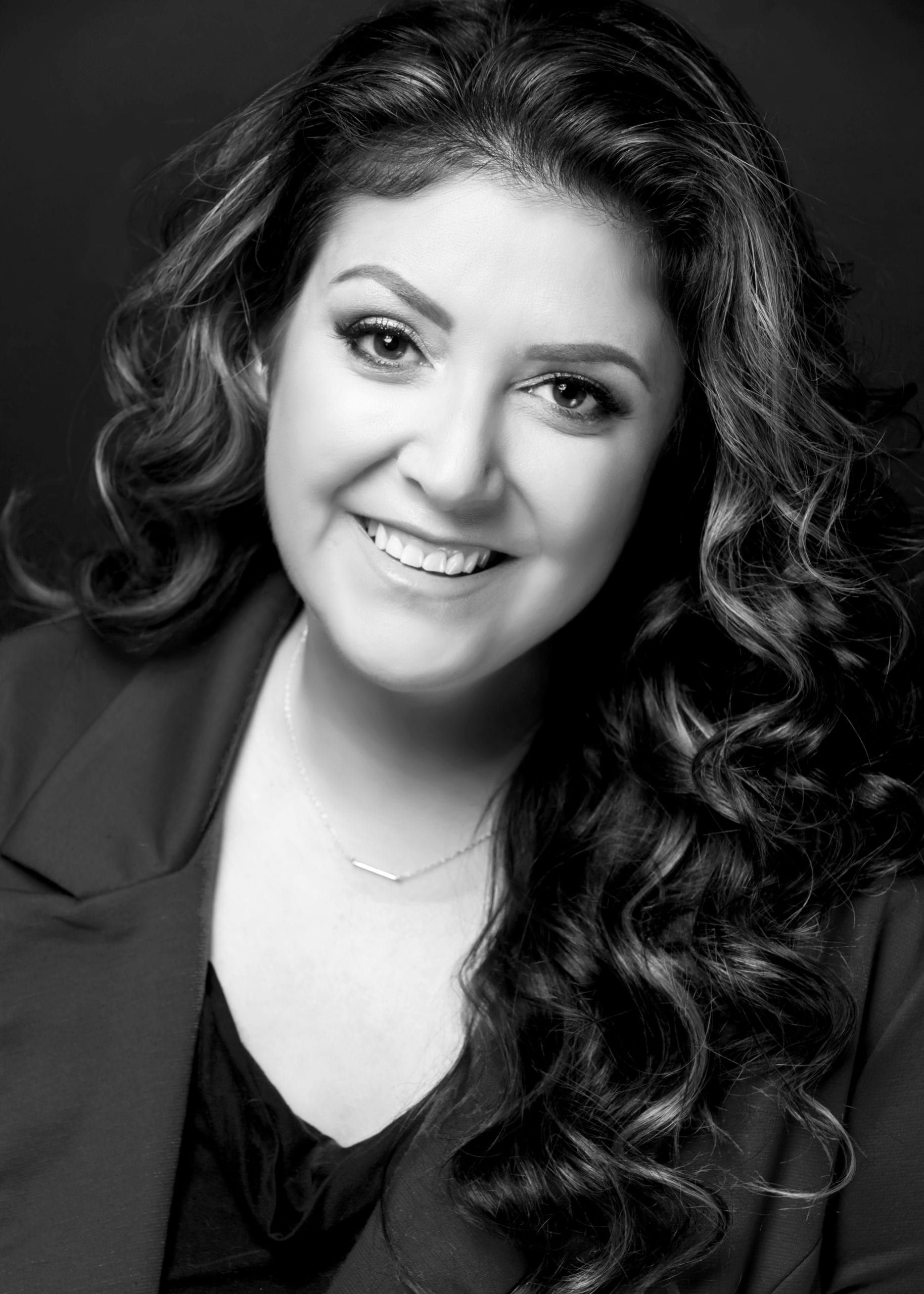 Smiling woman with curly hair, wearing a blazer and necklace.