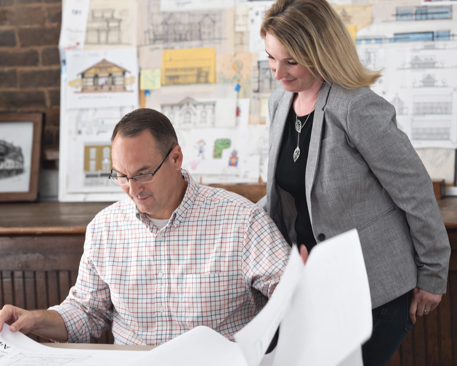 Man and woman reviewing architectural plans; workspace with sketches and brick wall.