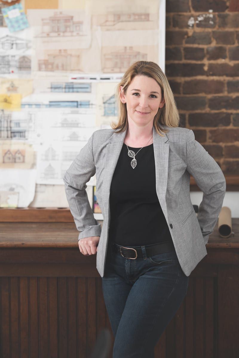 Woman in blazer, leaning on wooden counter.  Blonde hair, jeans, and black shirt.  Architectural drawings in background.