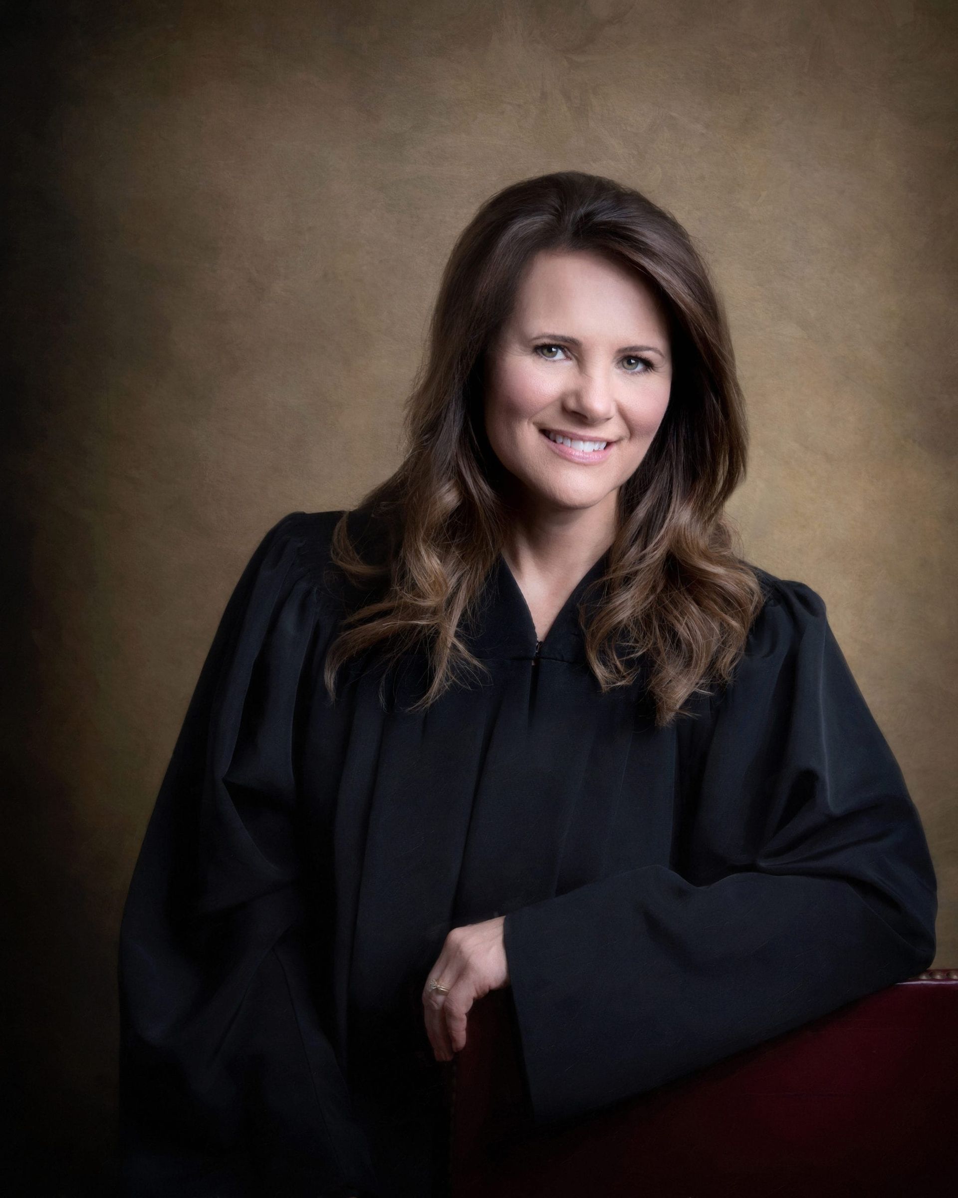 Woman in judge's robe smiles, leaning on a red chair; brown backdrop.