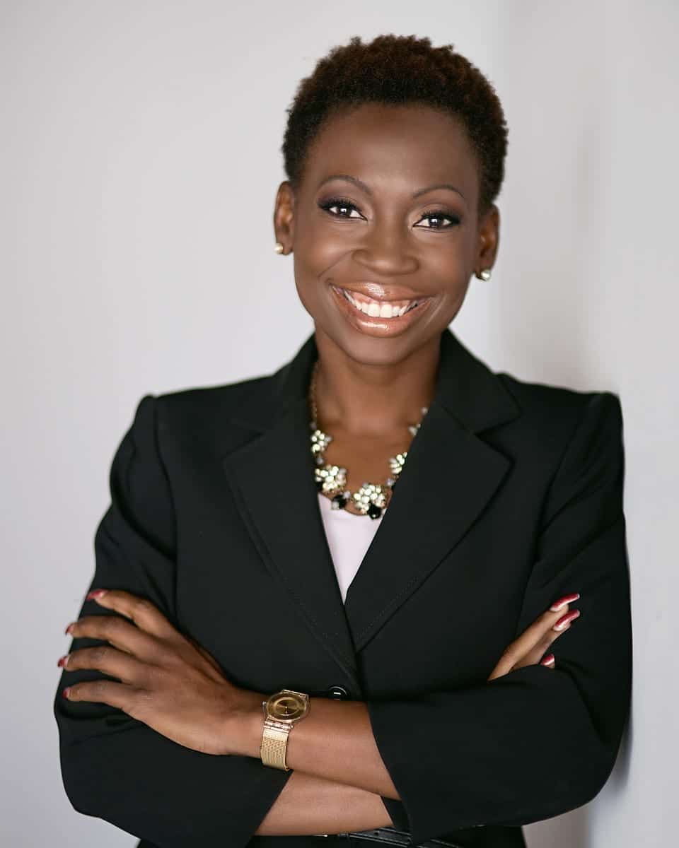 Woman with short hair in a black suit, arms crossed, smiling, wearing a gold watch and necklace.