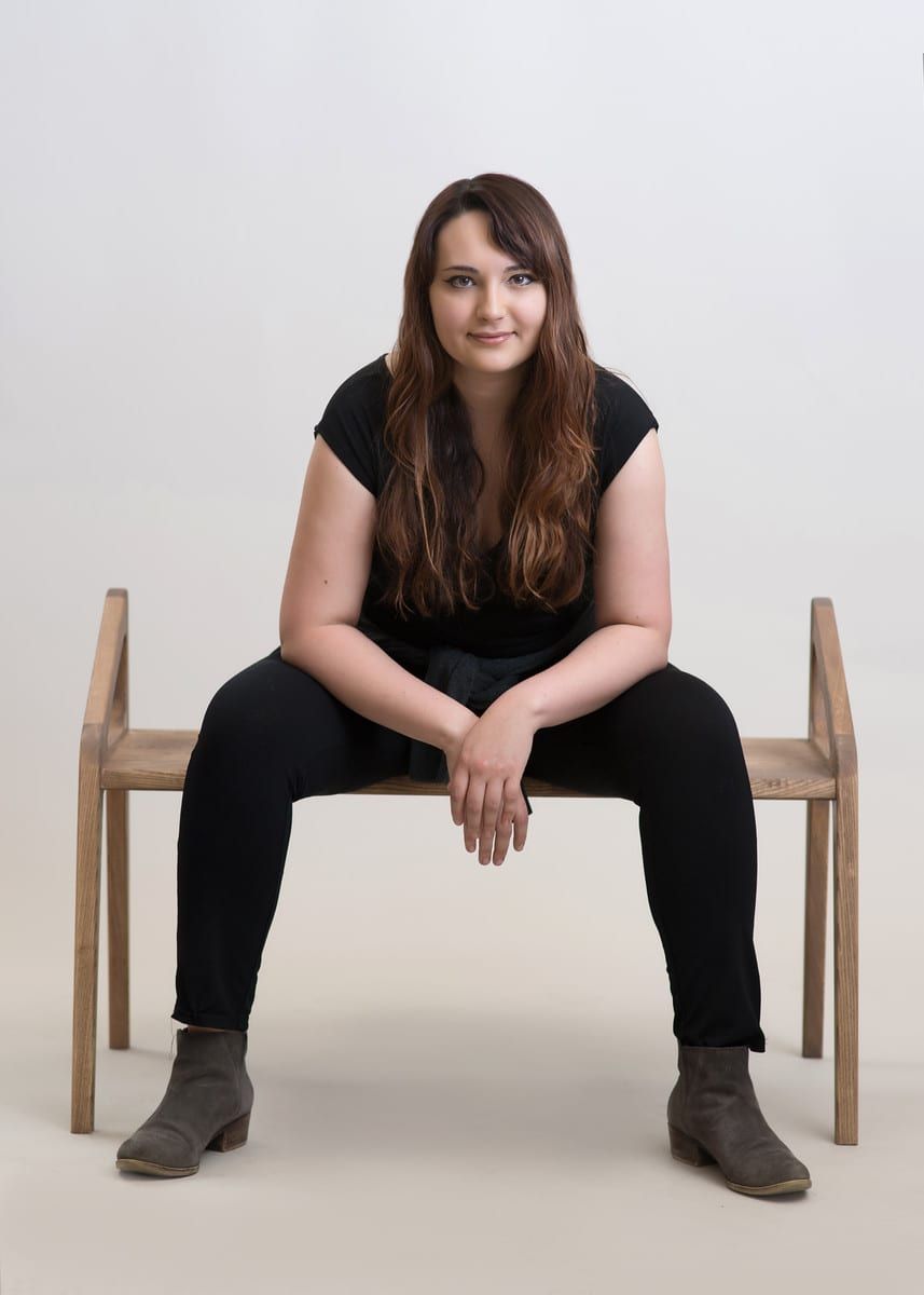 Woman in black outfit seated on wooden bench, legs spread, looking at the camera.
