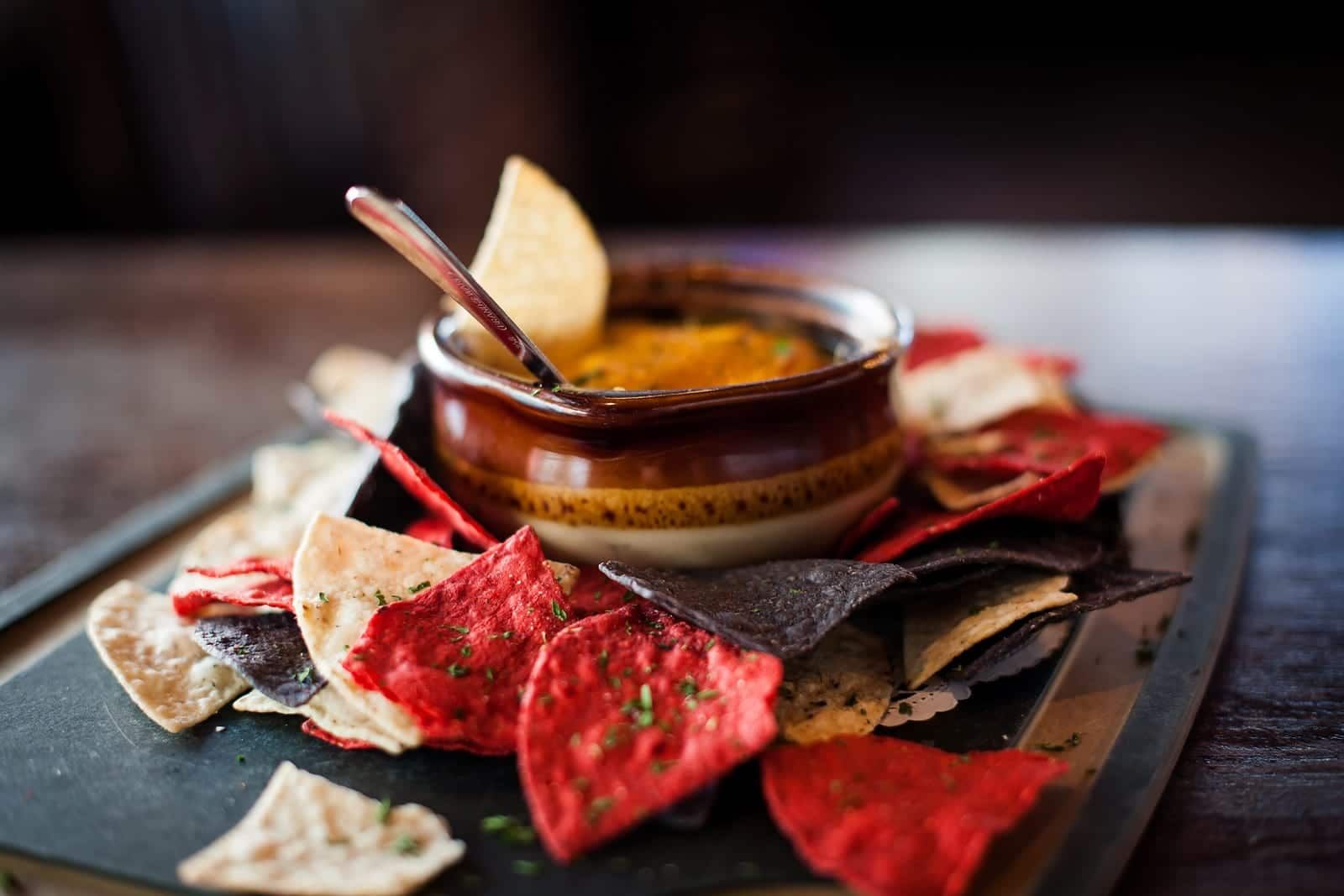 A small bowl of orange dip with a spoon, surrounded by red, blue, and white tortilla chips on a dark serving board.