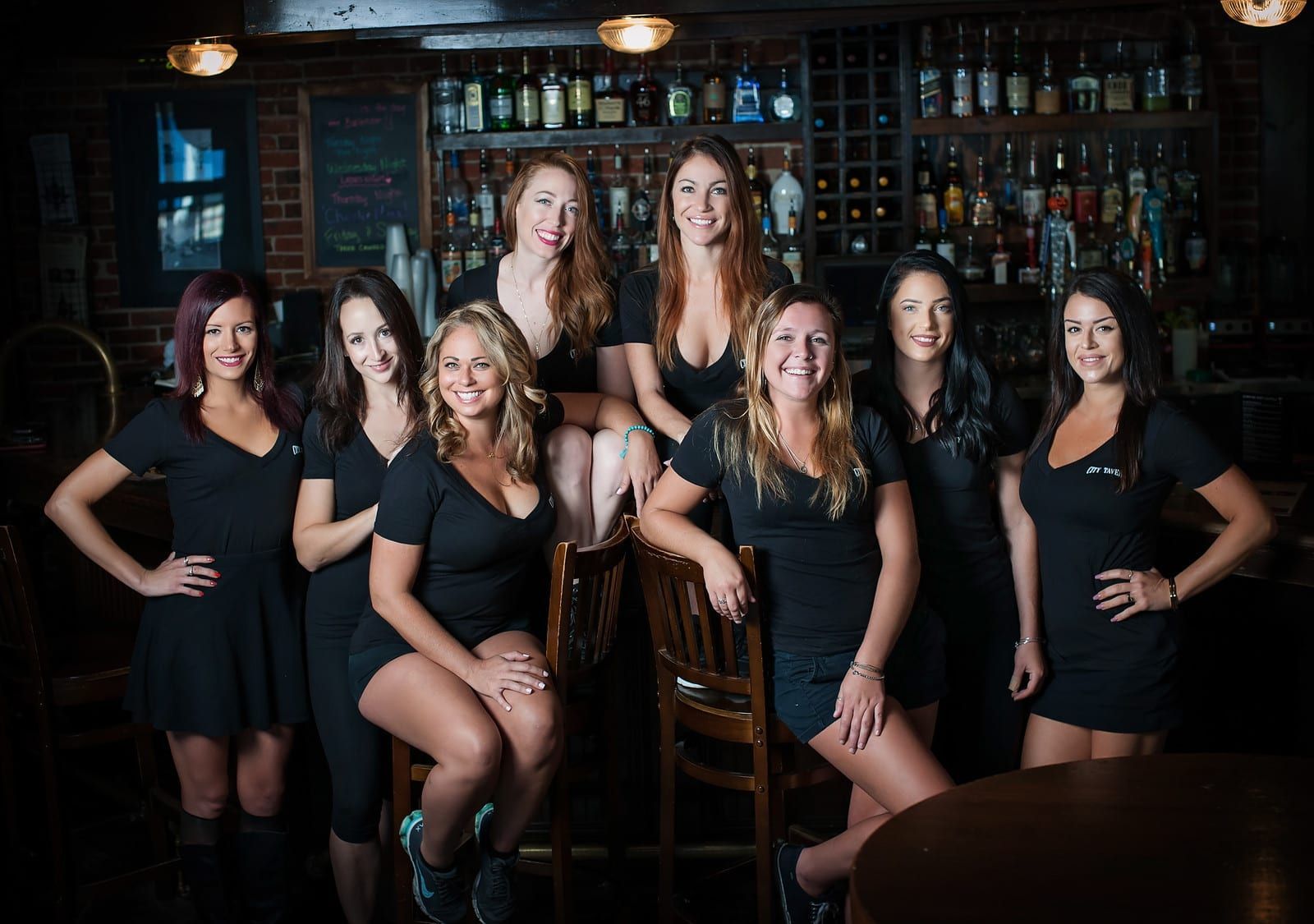 Group of women posing at a bar, wearing black, smiling, with a bar stocked with bottles in the background.