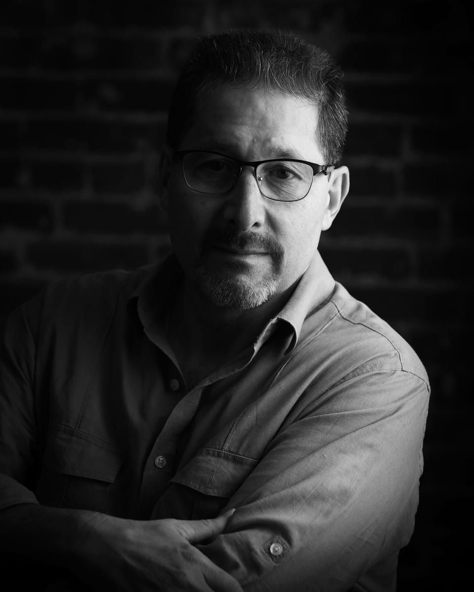Man with glasses, arms crossed, looking forward. Black and white portrait against brick wall.