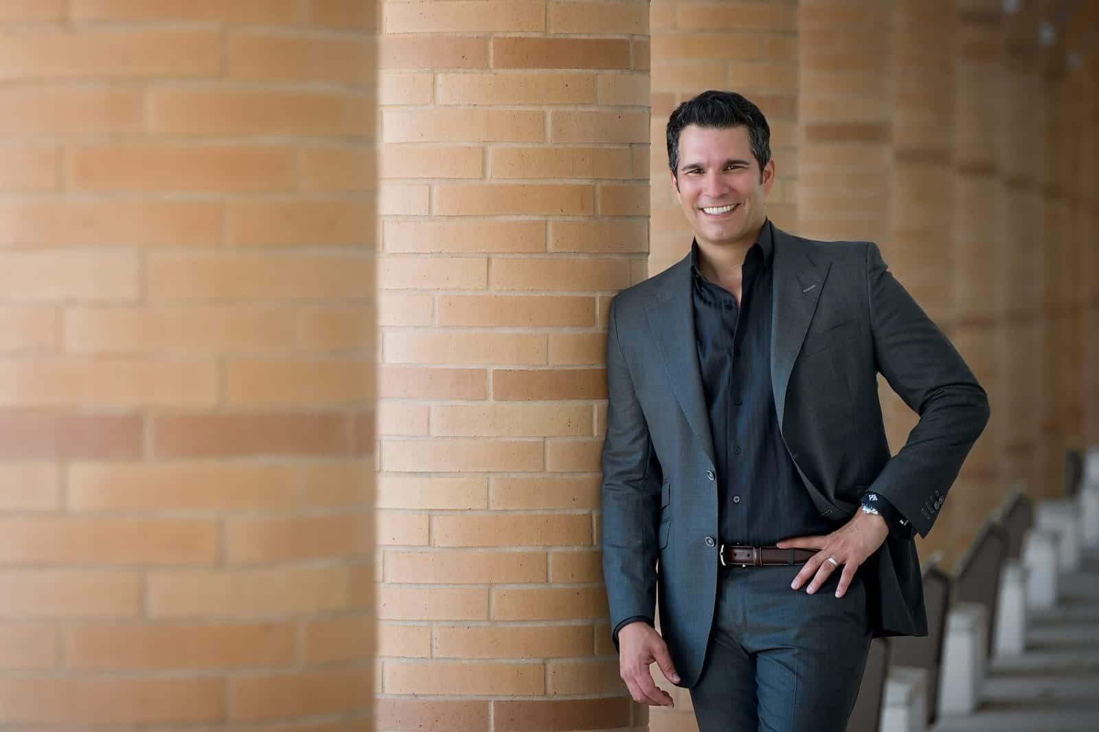 Man in a gray suit leans against a brick pillar, smiling.