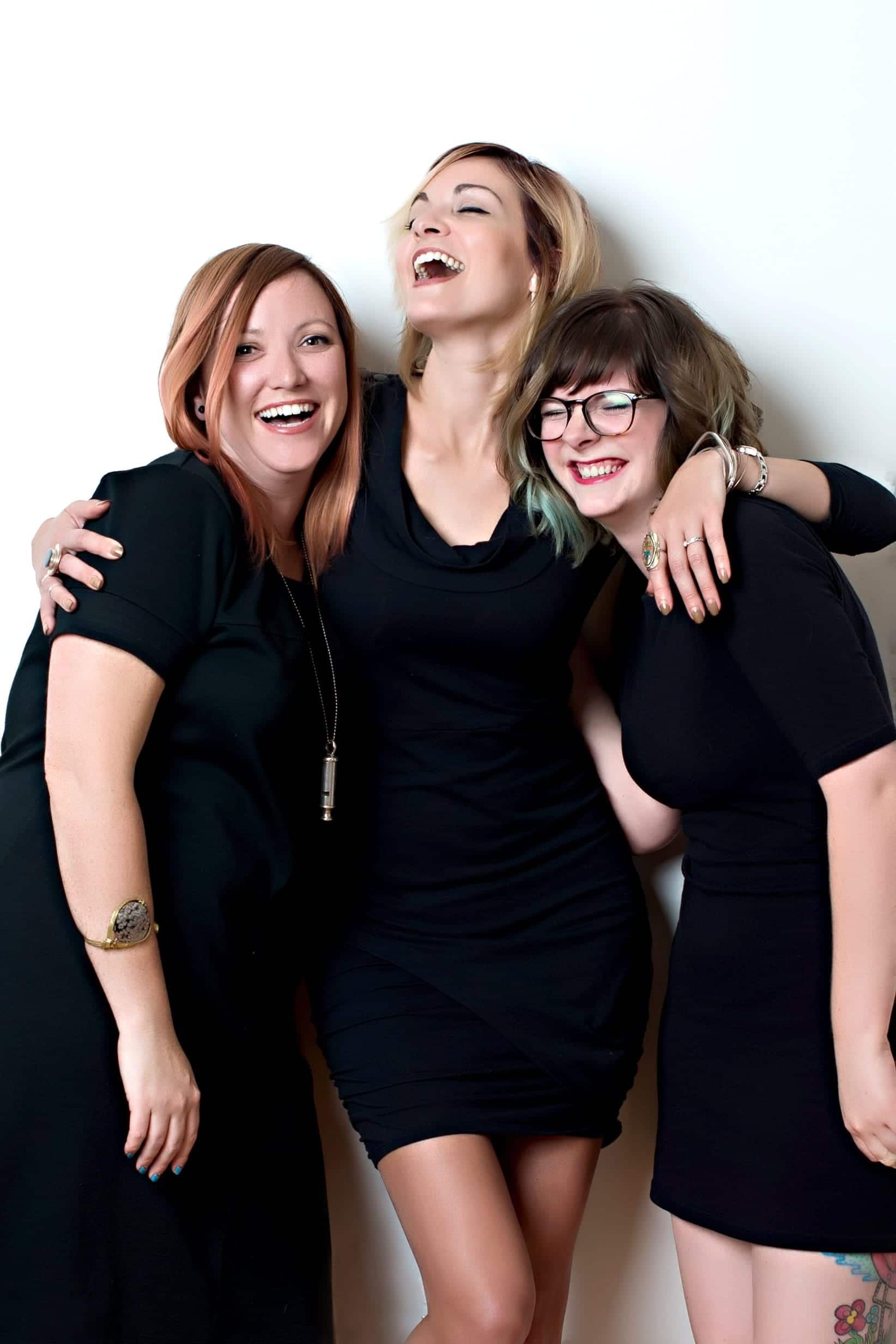 Three women in black dresses laughing, leaning against a white wall.