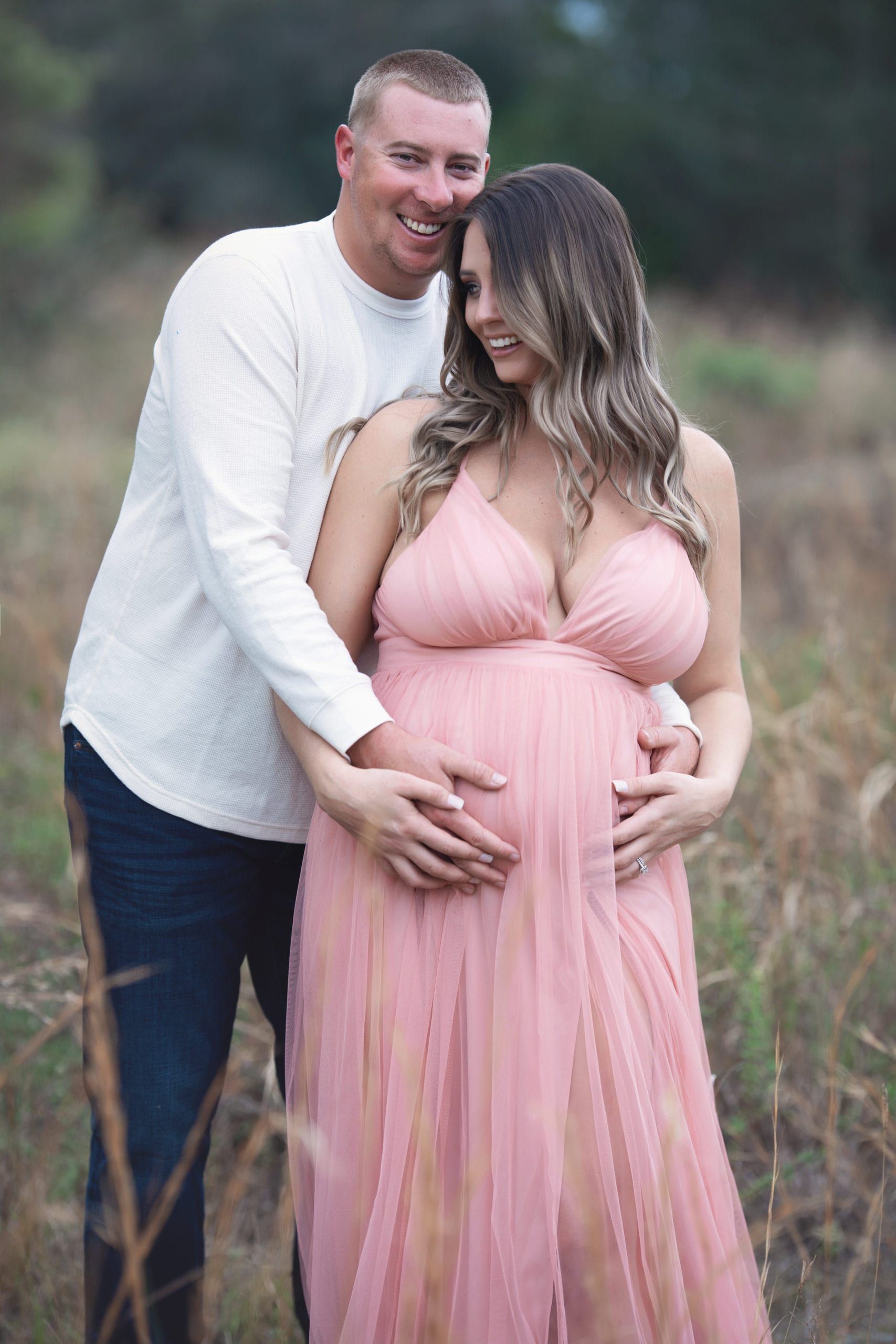 Couple in field, woman in pink dress cradles her pregnant belly, man hugs her, both smiling.