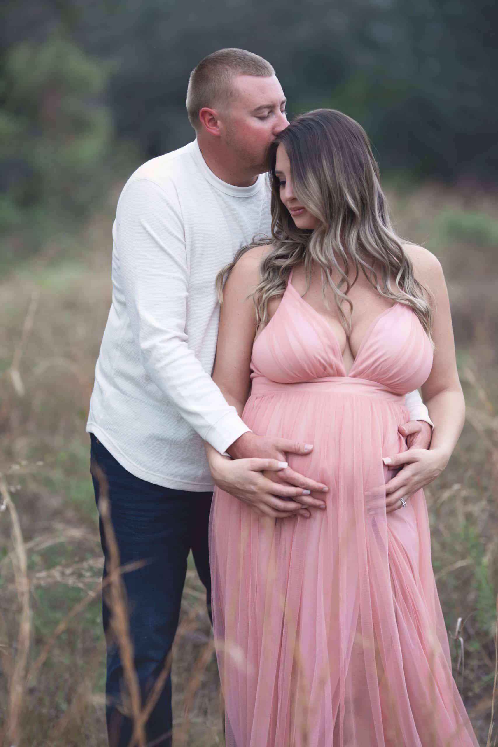 Pregnant woman in pink dress embraces man in field. He kisses her forehead and holds her belly.