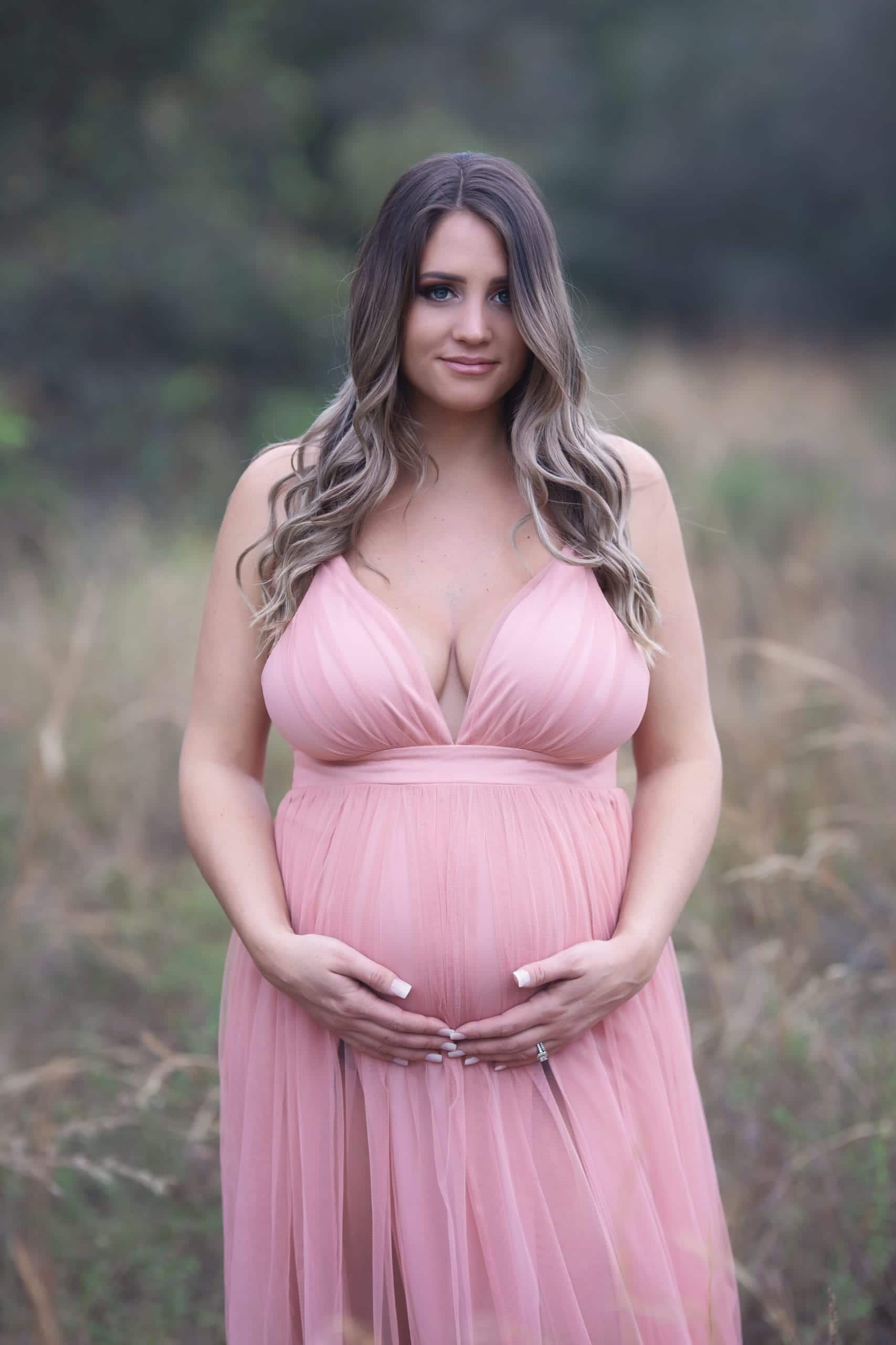 Pregnant woman in a pink dress, holding her belly, outdoors.
