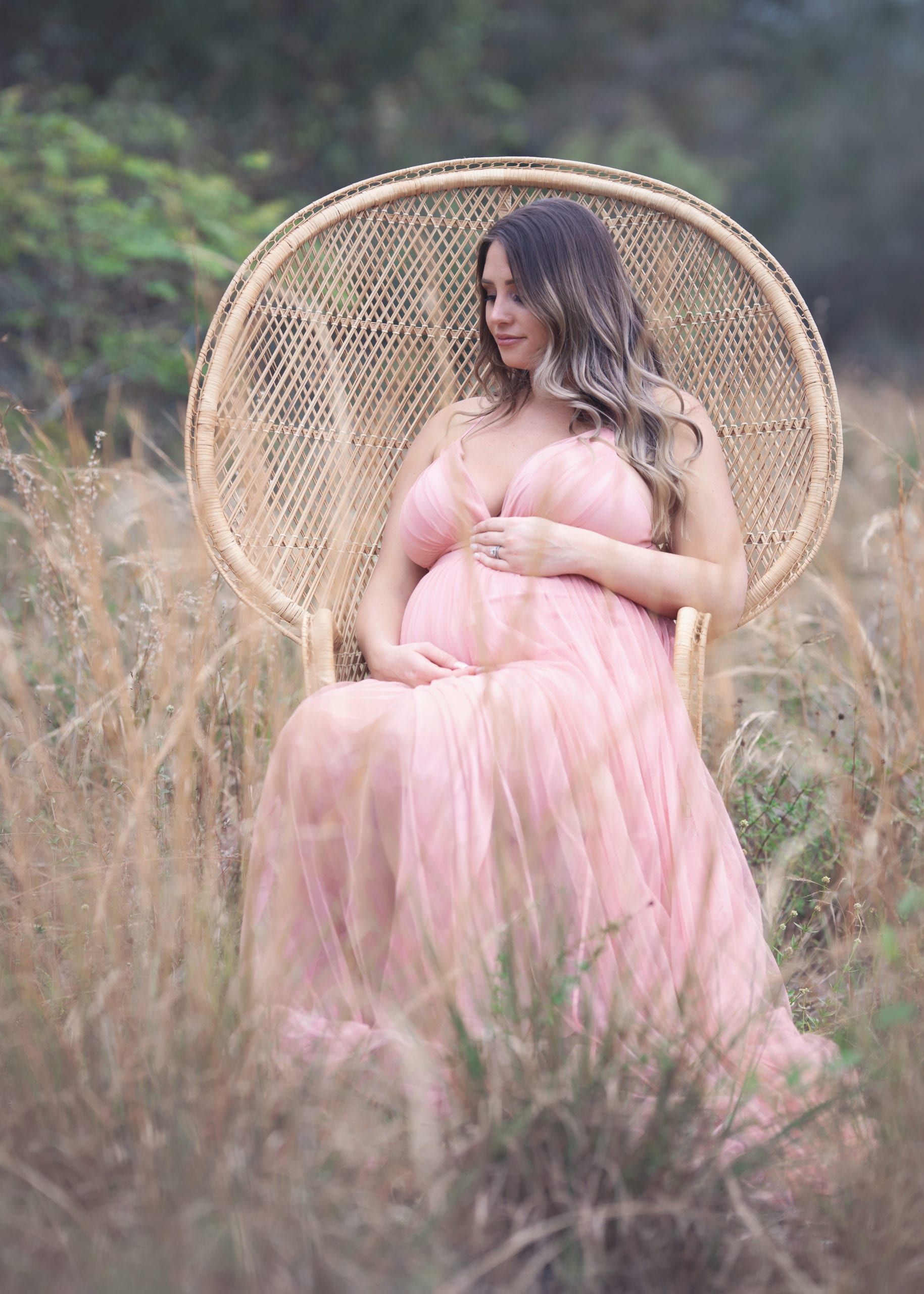 Pregnant woman in a pink dress sits in a peacock chair in a field.