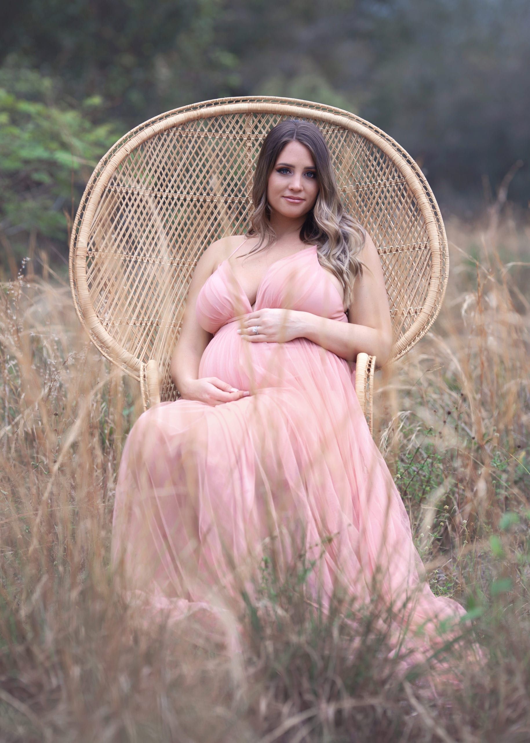 Pregnant woman in pink dress sits in woven peacock chair in field.