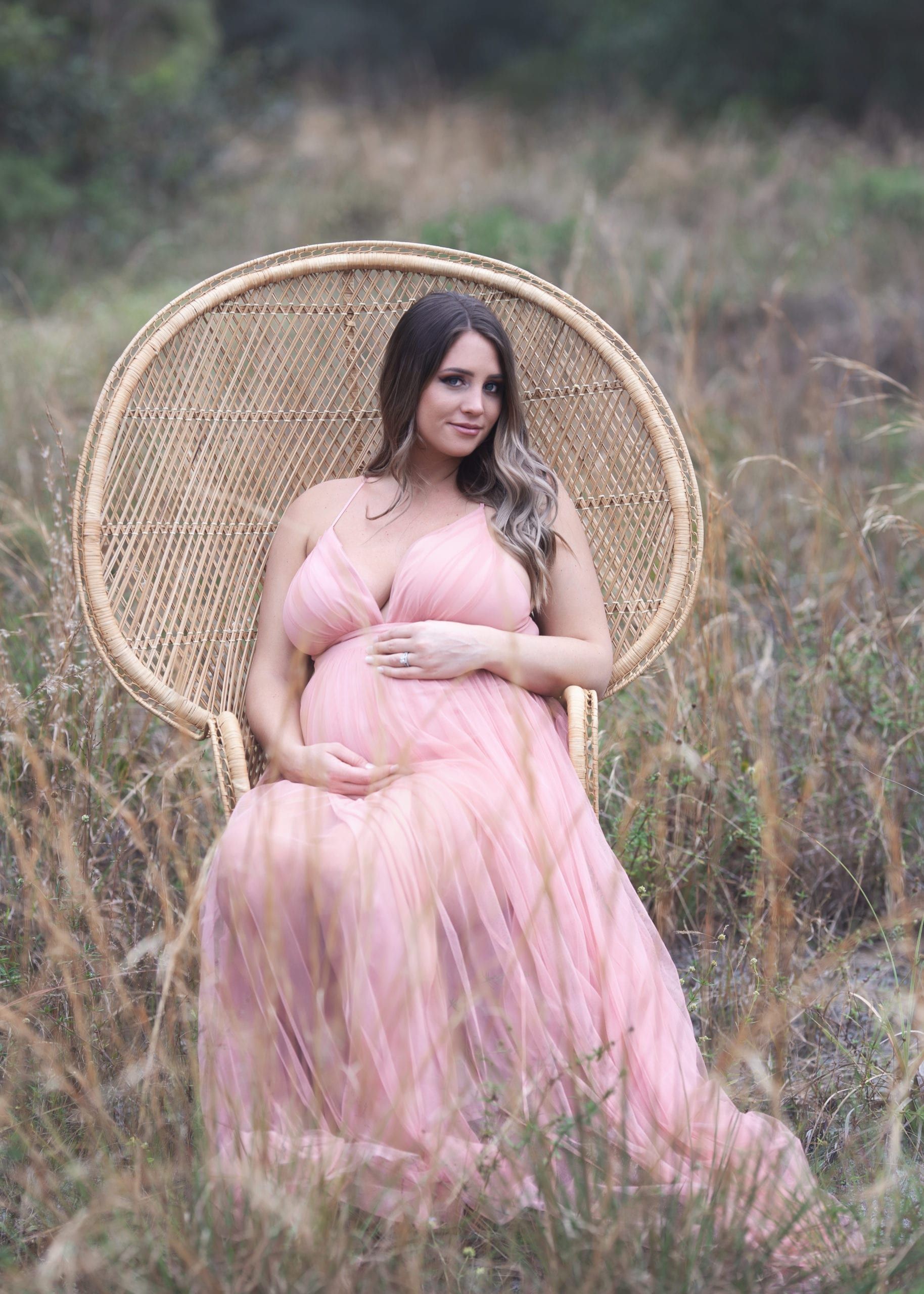 Pregnant woman in pink dress sitting in peacock chair outdoors.