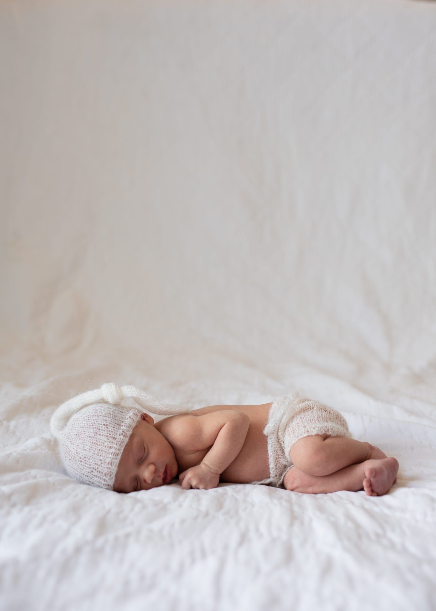 Newborn baby sleeping on white sheet, wearing knitted hat and diaper cover.