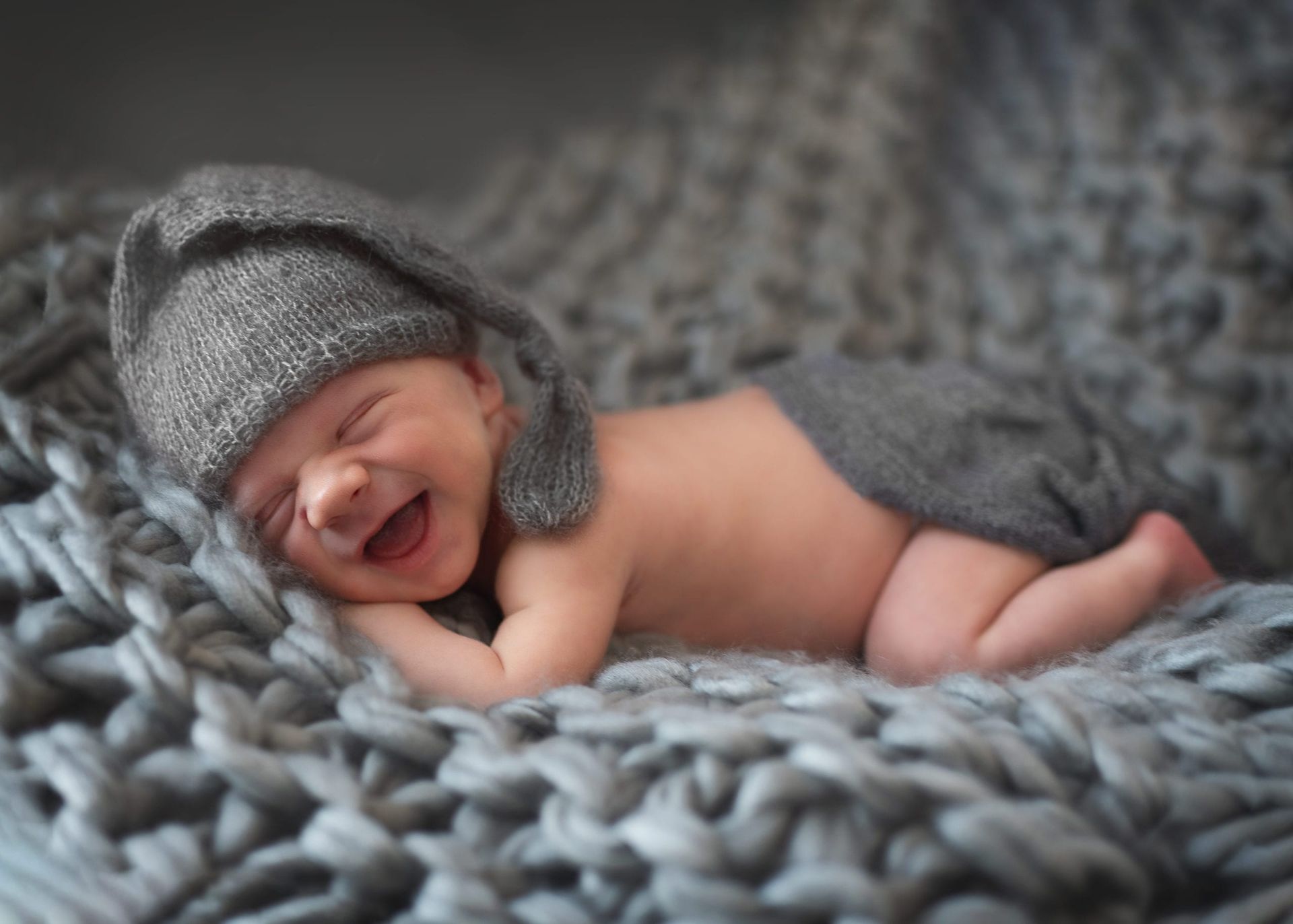 Smiling newborn wearing a gray hat and diaper, lying on a gray knitted blanket.