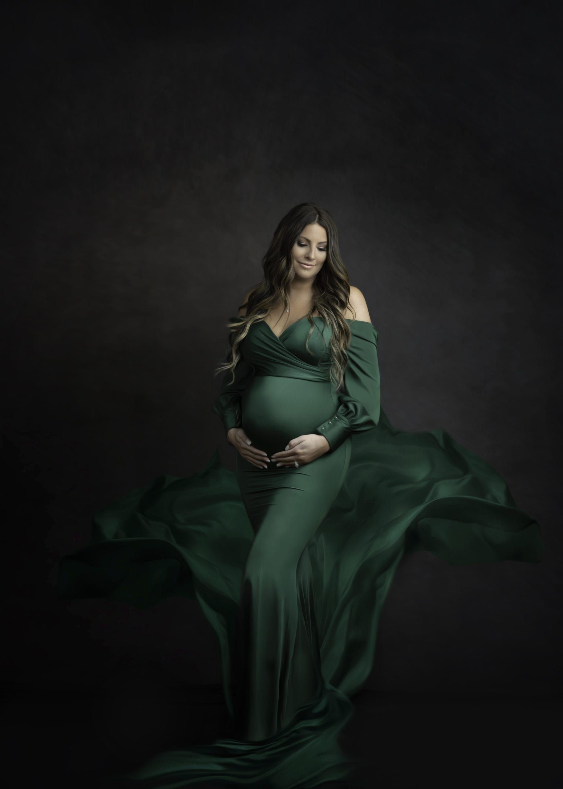 Pregnant woman in emerald green gown cradles belly, smiling against dark backdrop.
