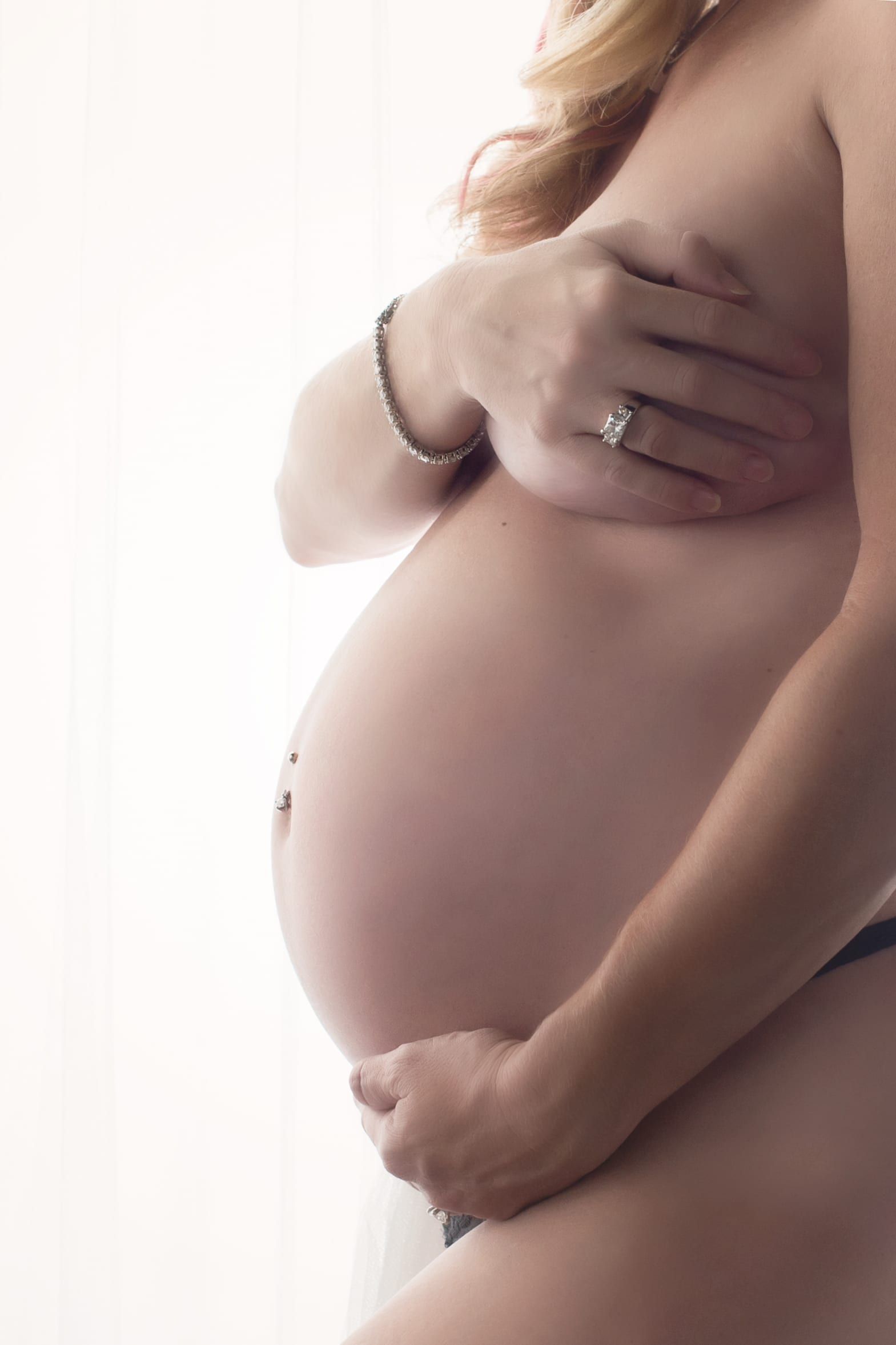 Pregnant woman, belly prominent, tenderly cradled by her hands, lit by soft window light.