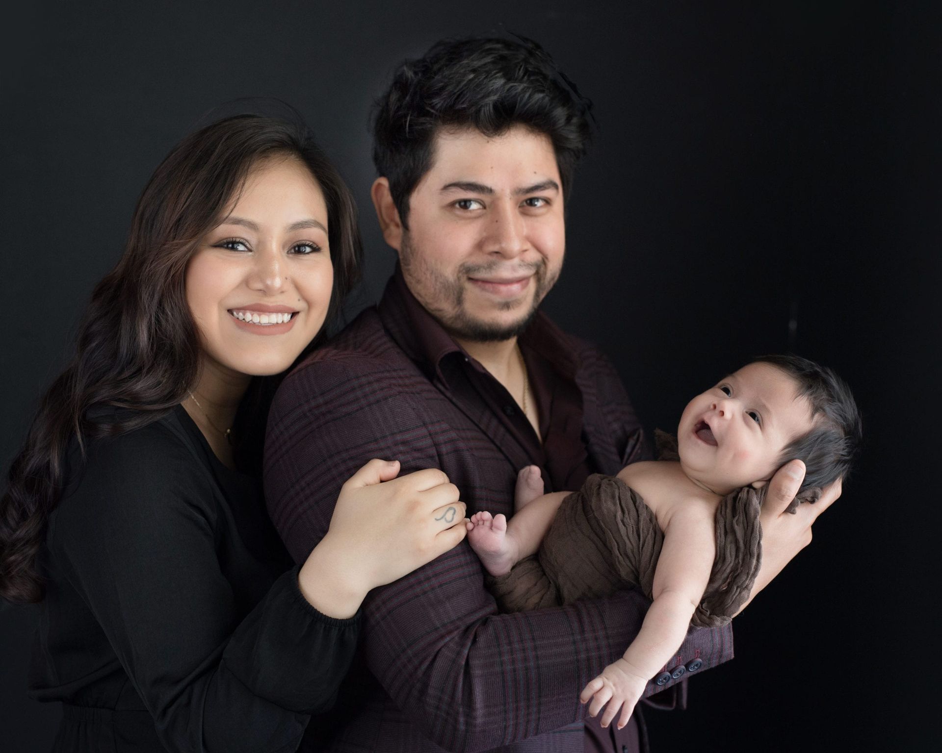 Smiling family of three poses against a black background; parents hold a newborn baby.